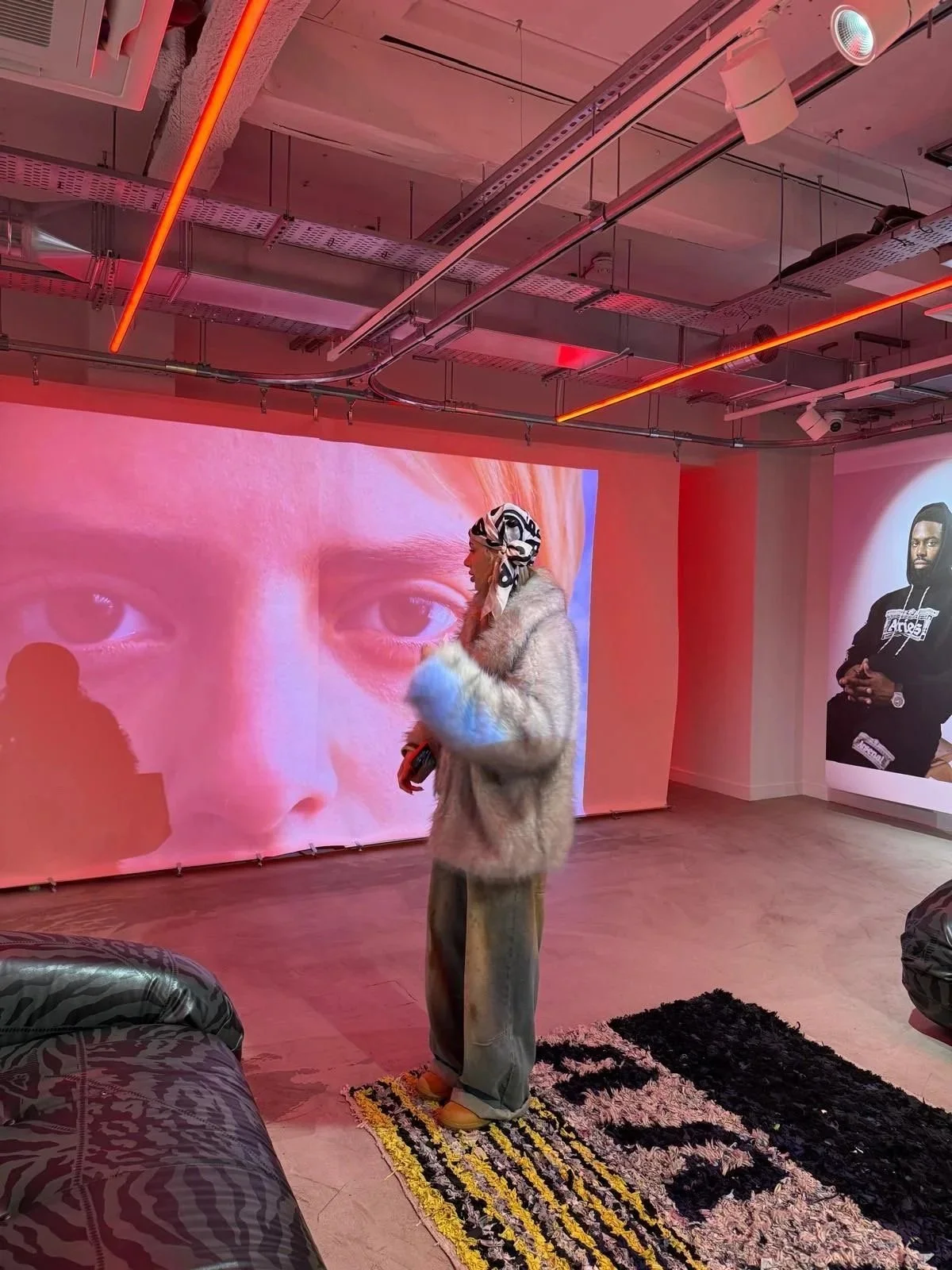 A person dressed in a fur coat, loose pants, and a headscarf stands in an art gallery with contemporary artwork and large video projections on the walls. The gallery has exposed ceiling pipes and colorful lighting.