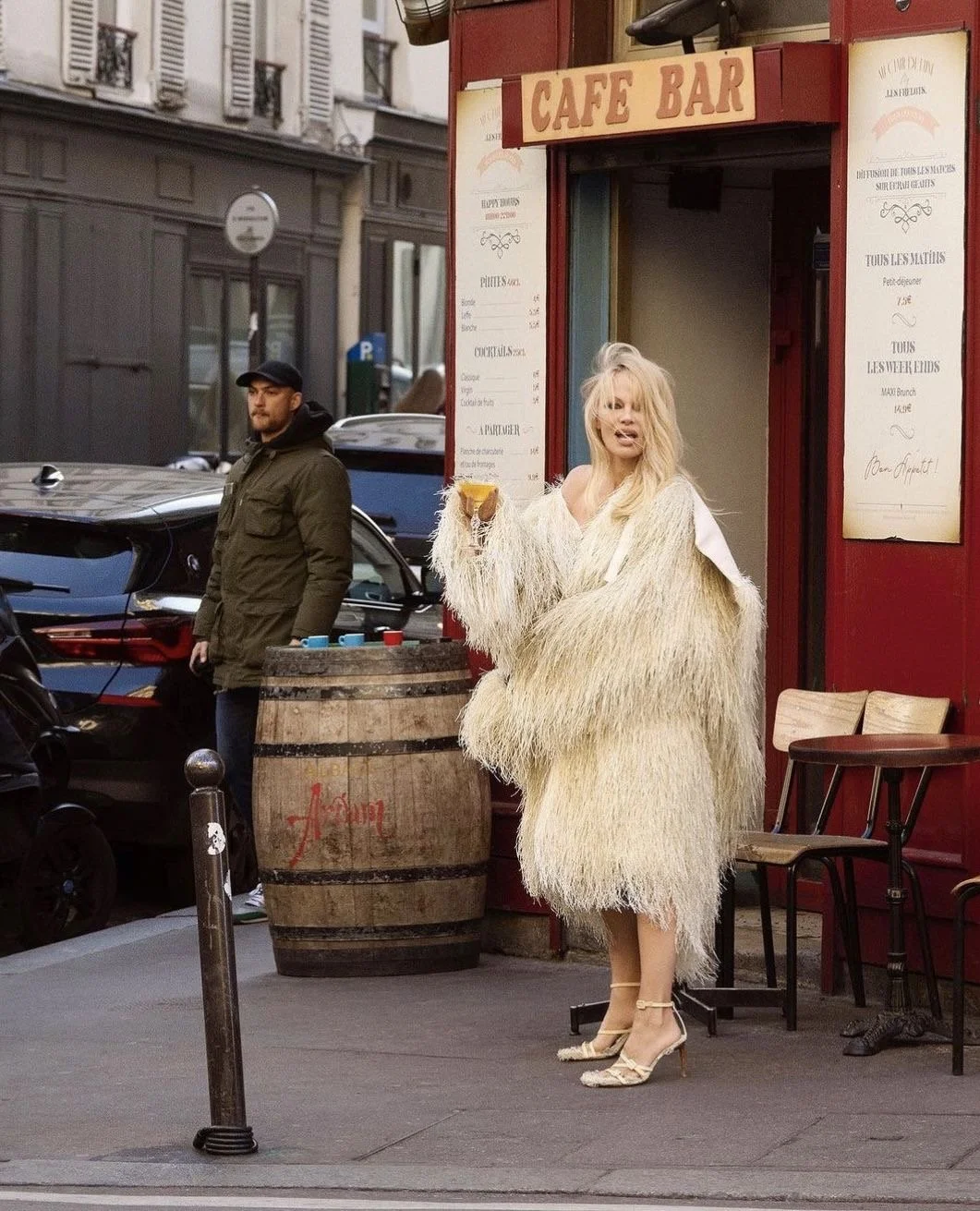 Woman in a large, shaggy, cream-colored coat and high heels standing outside a café bar, holding a drink, with a man standing nearby and cars parked on the street in the background.