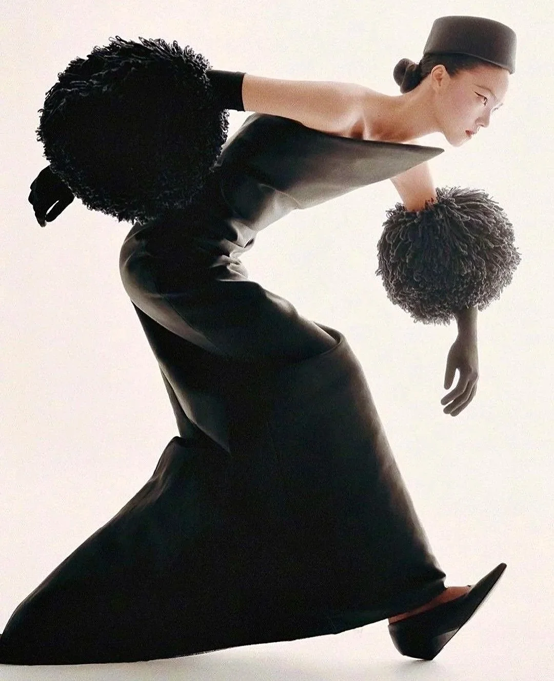 Fashion model wearing black leather outfit with large black gloves and a hat, posing against a white background.