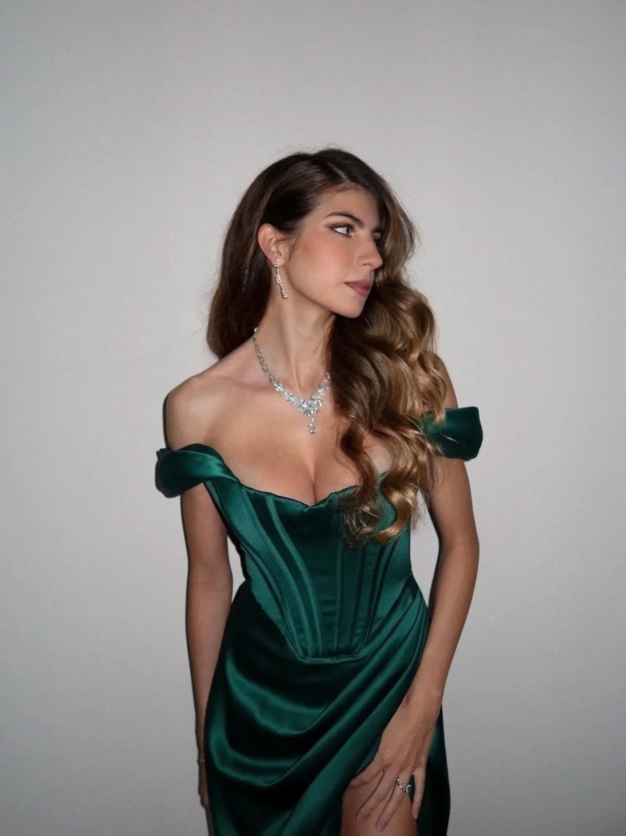 A woman with long, wavy brown hair wearing an emerald green satin dress with off-shoulder ruffled sleeves, accessorized with diamond jewelry, standing against a plain light gray background.