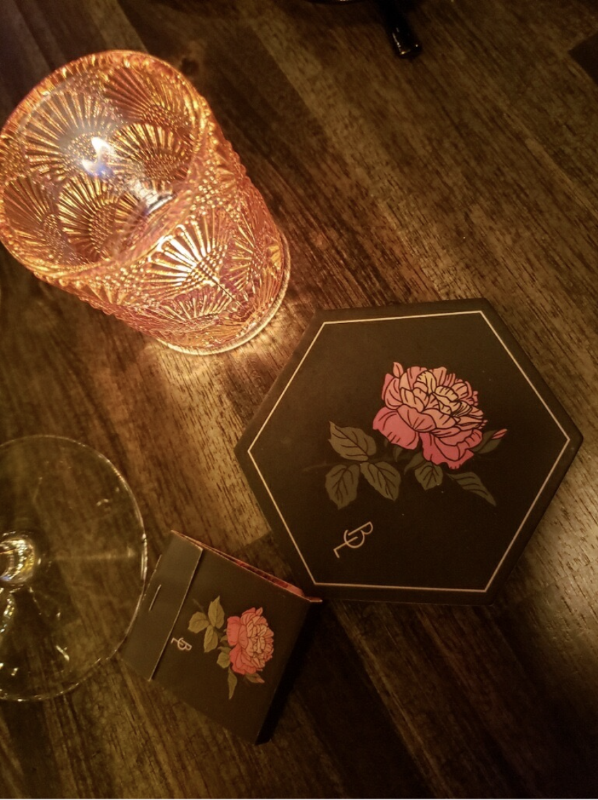 A pink patterned glass candle holder, a box with a pink rose illustration, and an empty wine glass on a wooden table with a dark heart-shaped box featuring a pink rose design.