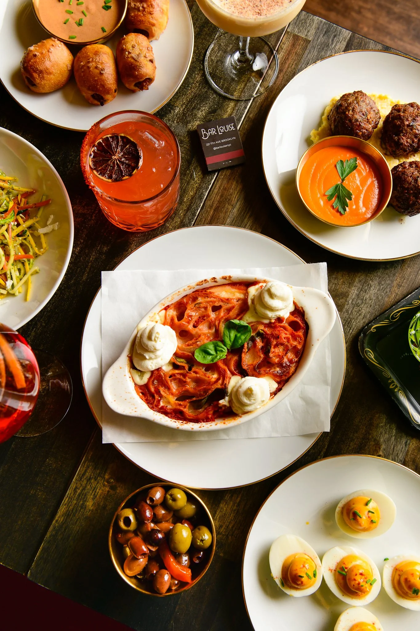 Assorted dishes including deviled eggs, meatballs, pasta with tomato sauce, egg drop soup with chopped herbs, and a drink with a citrus slice and ice on a dark wooden table.