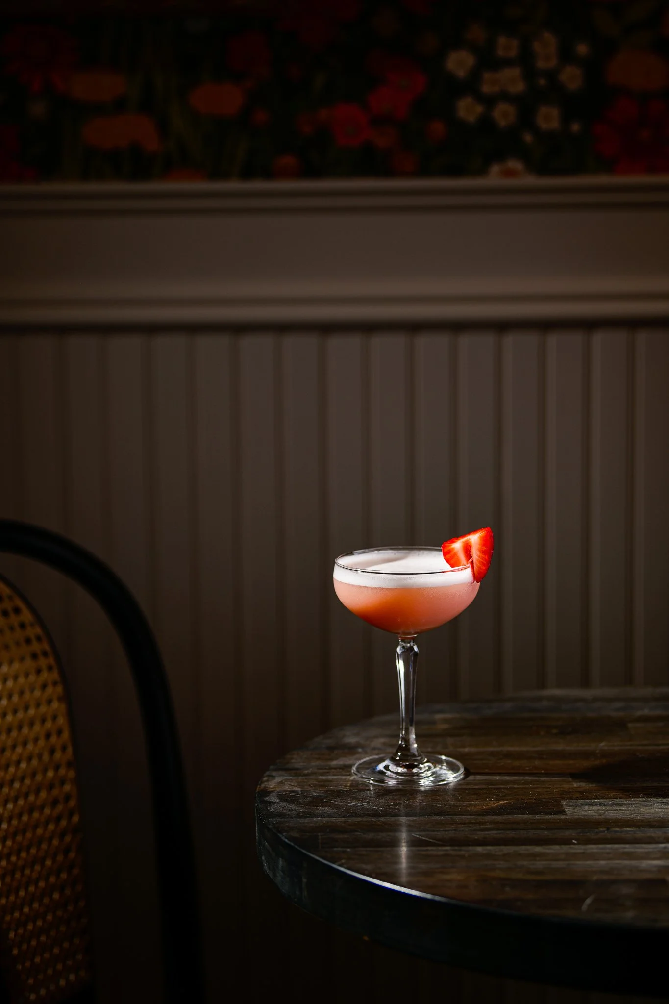 A pink cocktail with a strawberry garnish on a wine glass, on a dark wooden table.