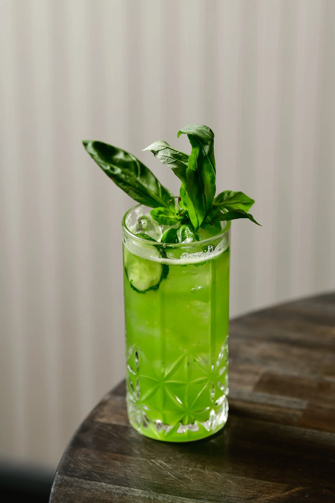 A tall, clear glass filled with bright green liquid and ice, garnished with fresh green basil leaves, placed on a wooden surface against a light-colored background.