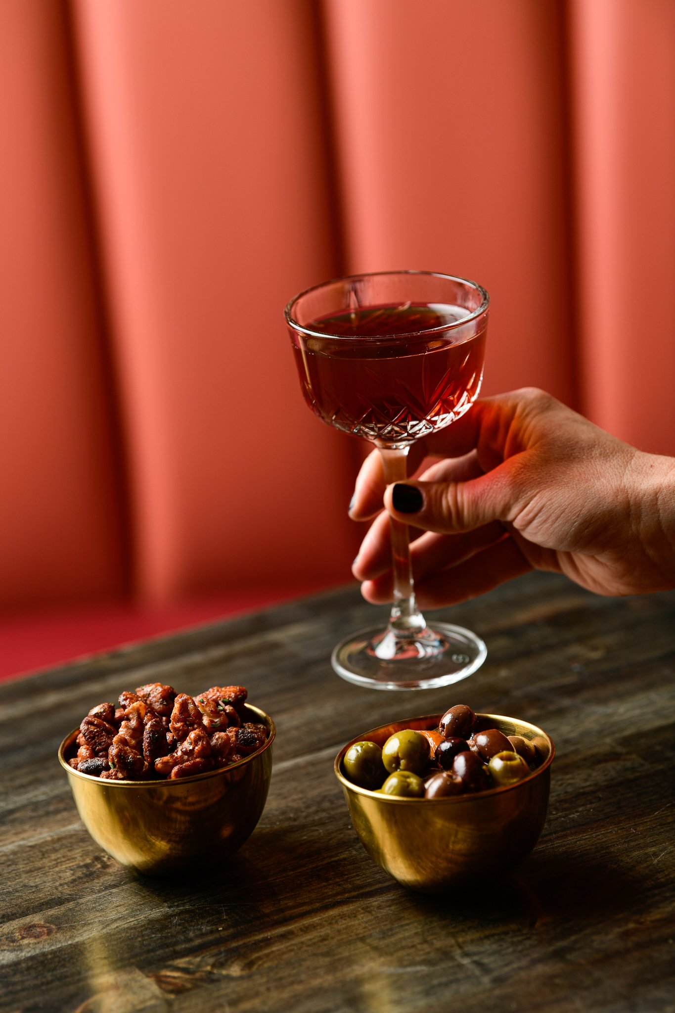 A hand holding a glass of red wine against a pink background, with bowls of mixed olives and seasoned nuts on a wooden table in the foreground.