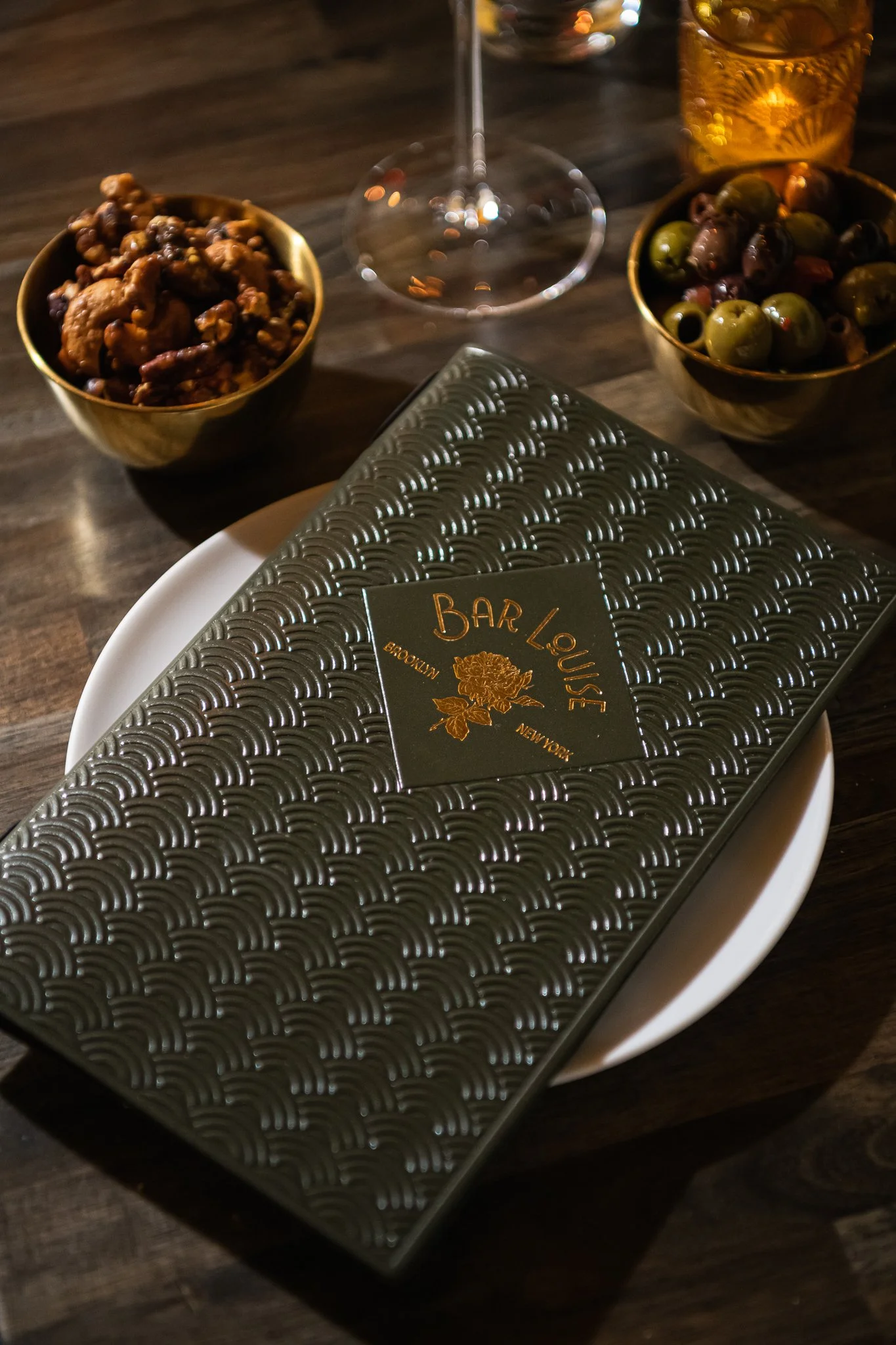 A black and gold patterned menu on a white plate, with bowls of mixed nuts and green olives nearby, set on a dark wooden table.