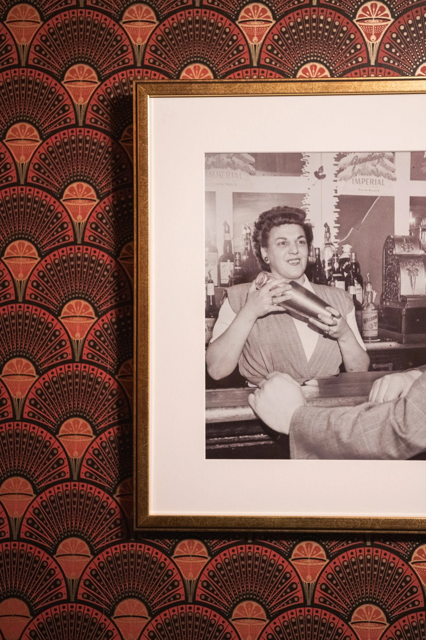 A black and white vintage photograph of a woman behind a bar, smiling and holding a cocktail shaker, with bottles and bar accessories in the background.