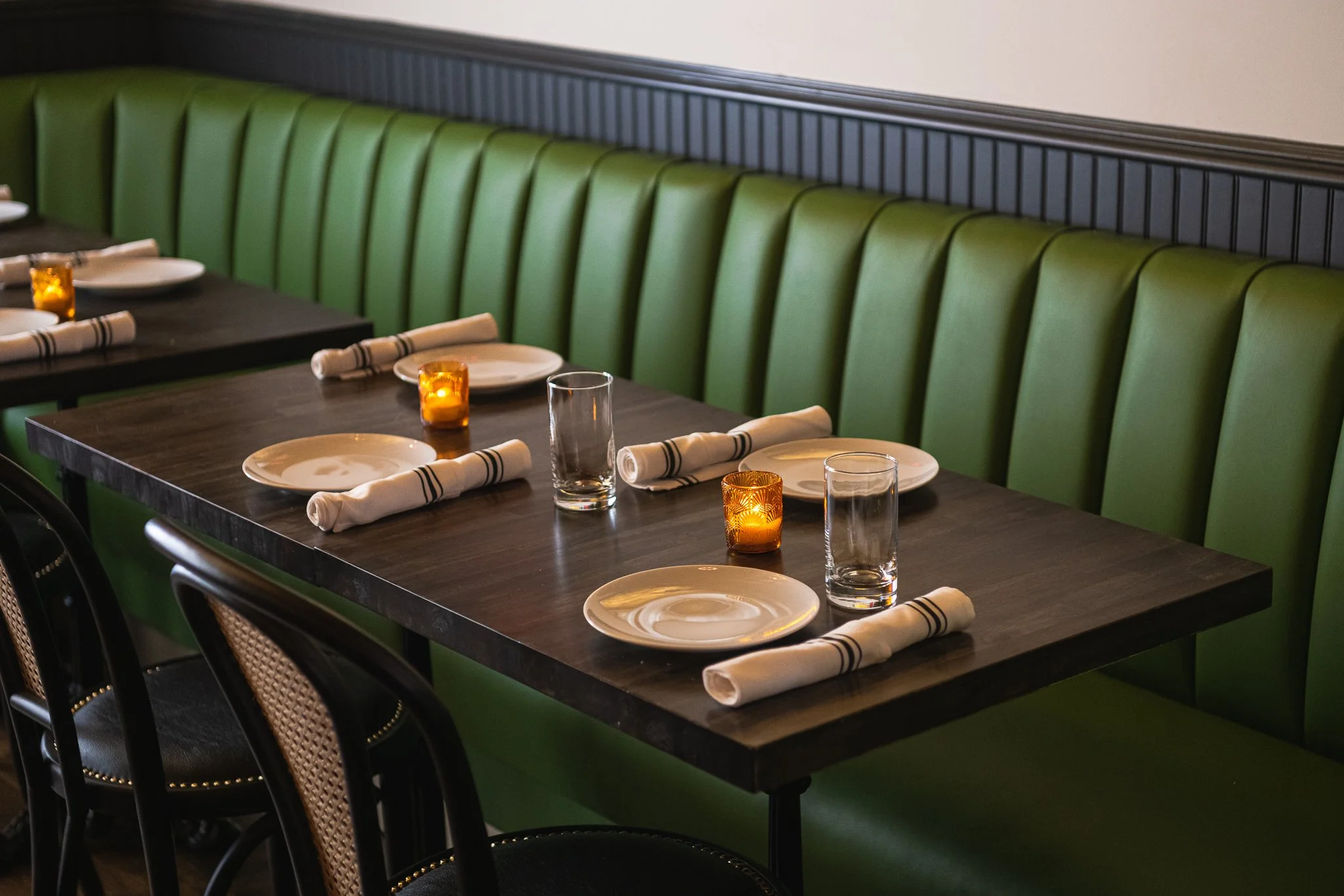 A restaurant table set with white plates, rolled napkins, glasses, and small orange candle holders, with a green cushioned bench and black chairs along a dark wooden table.