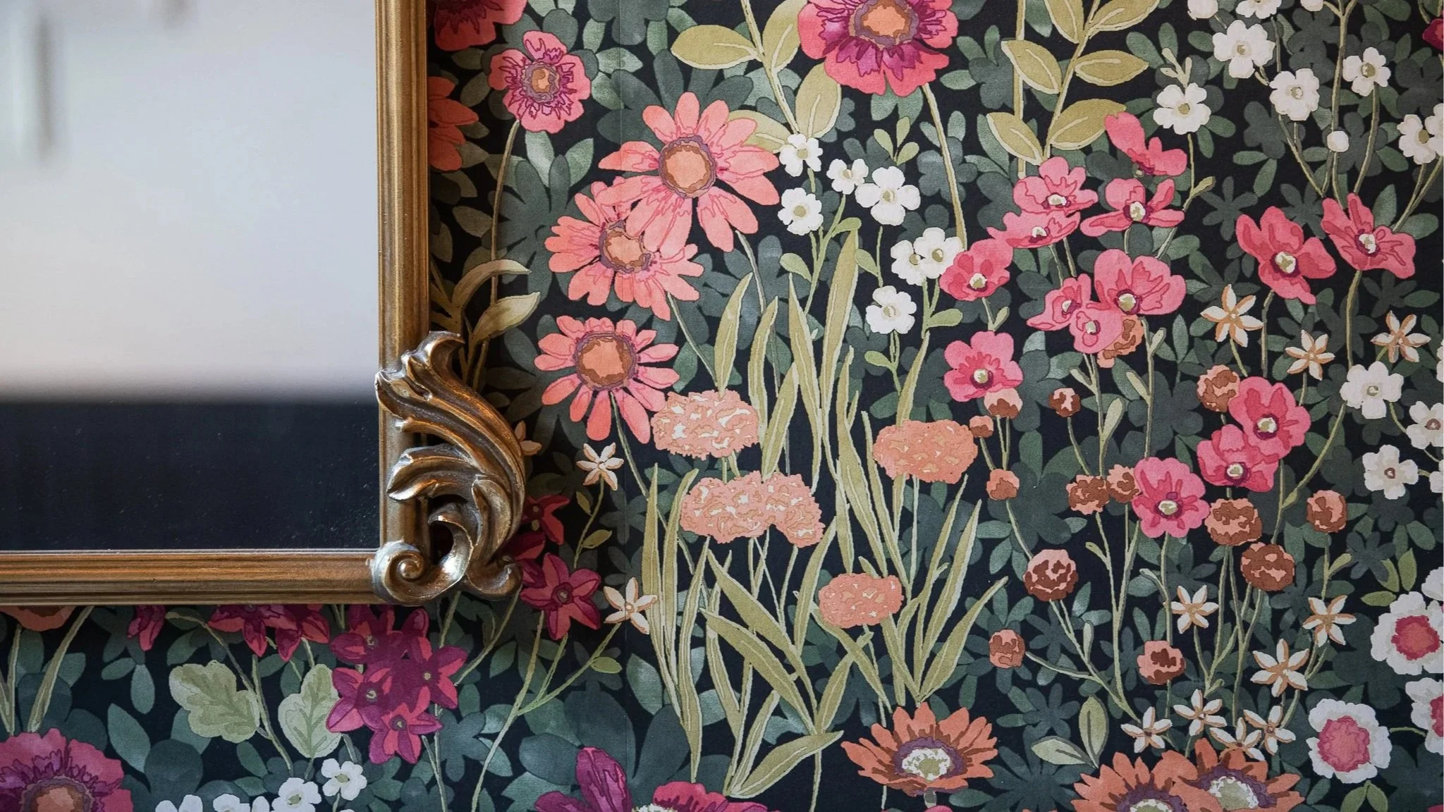 Close-up of a gold-framed mirror with a floral-patterned wallpaper background featuring pink, white, and brown flowers and green leaves.