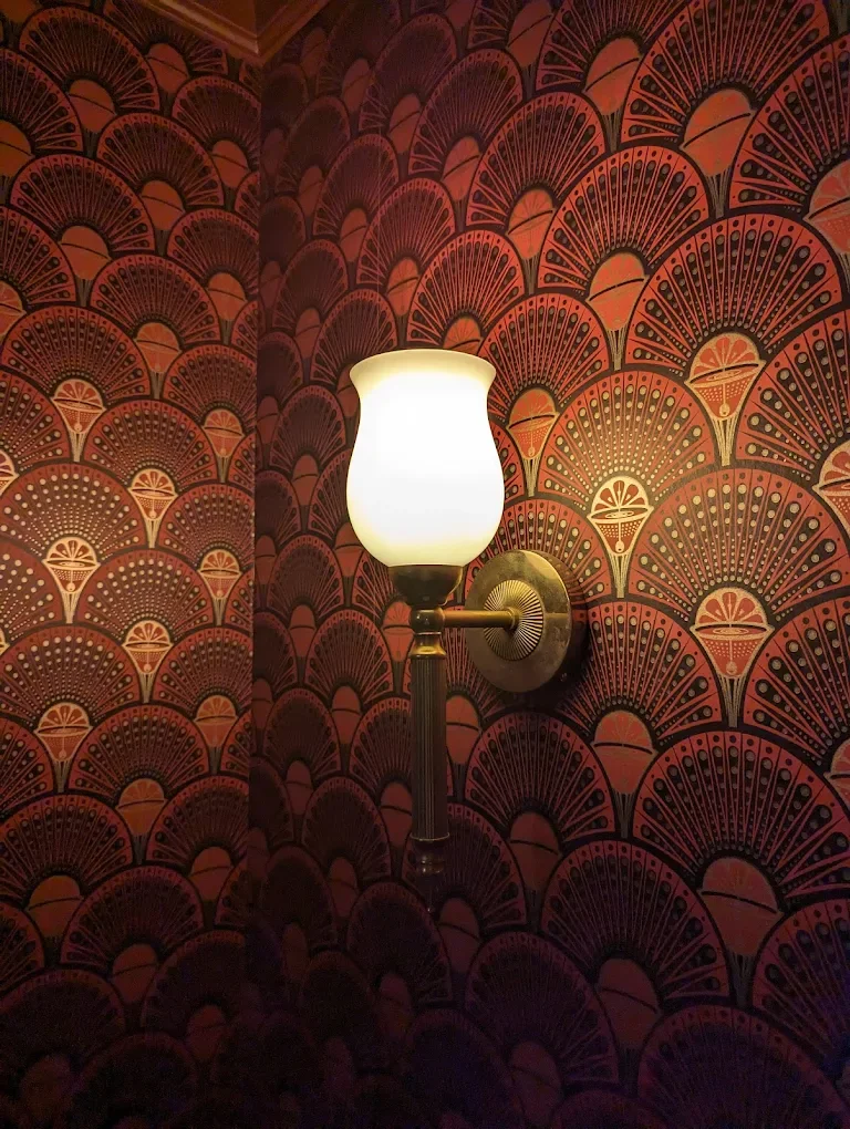 A wall-mounted lamp with a frosted glass shade and brass fixture, set against a decorative wallpaper with a fan pattern in red and gold colors.