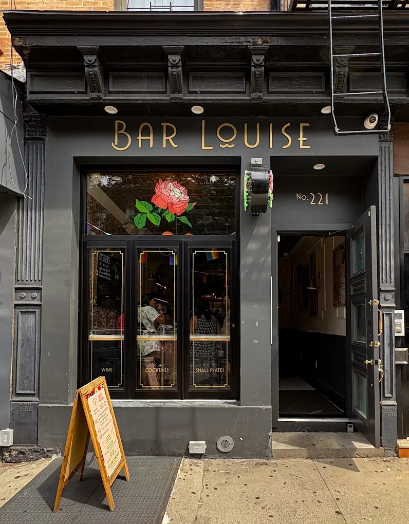 Exterior of a bar named 'Bar Louise' with black storefront, large window with a painted pink rose, and an open door. A small wooden menu stand is on the sidewalk outside.