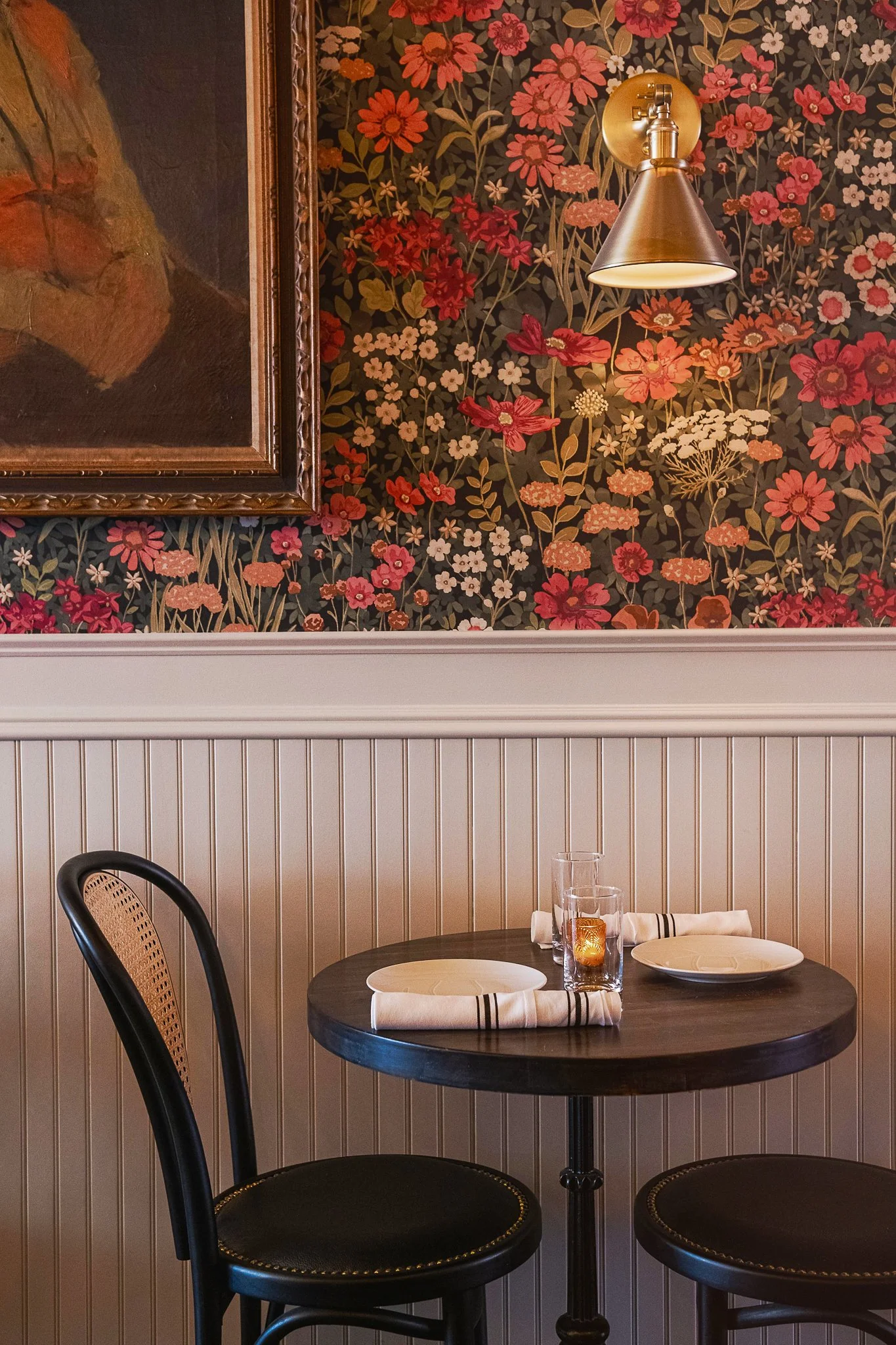 A small round dining table set for two with two black chairs, white plates, napkins, a glass with a candle, and a white cloth napkin in a restaurant with floral wallpaper and framed artwork.