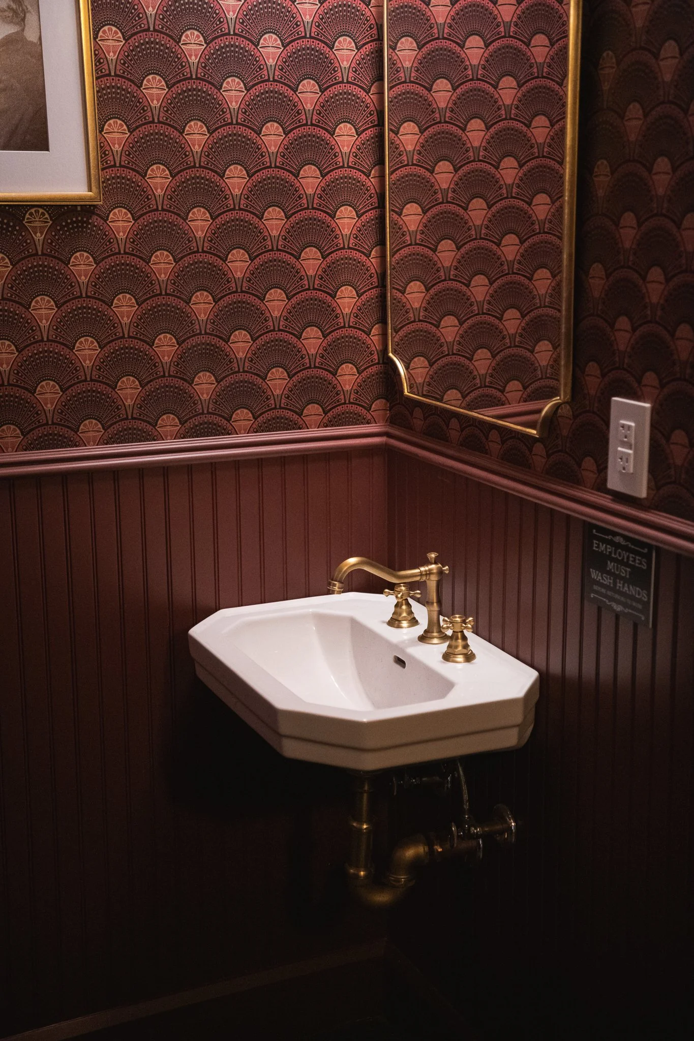 A bathroom with dark red and black art deco patterned wallpaper, a gold-framed mirror, a white wall-mounted sink with gold faucets, and a black wall with wainscoting. There is a smoke detector and a small section of a framed picture in the upper left corner.