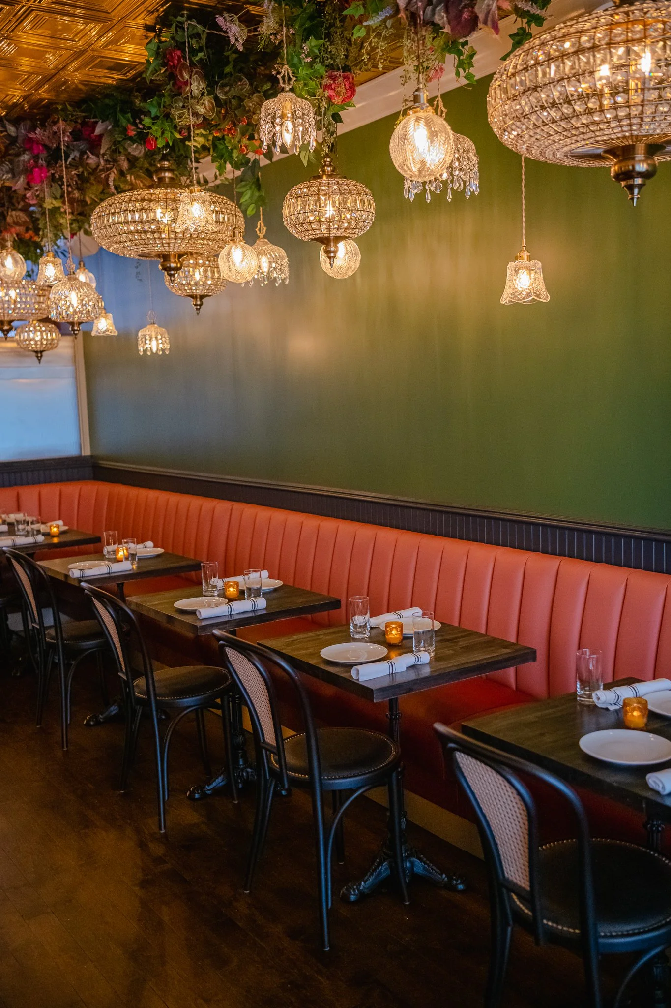Elegant restaurant interior with a green wall, pink velvet banquette seating, dark wooden tables, and a row of vintage-style chandeliers hanging from a floral ceiling.