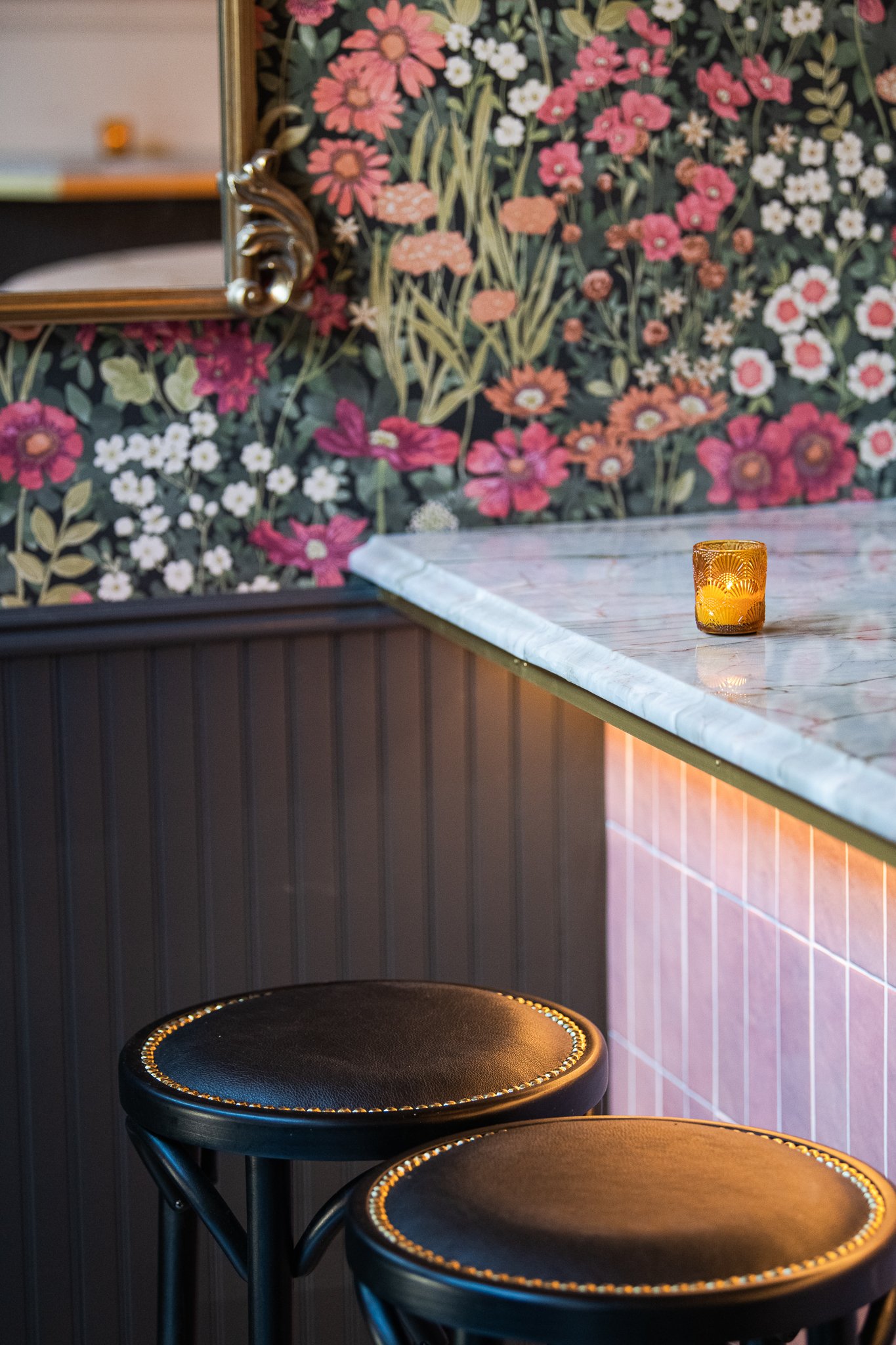 Three black chairs with gold trim positioned against a marble counter, with a floral-patterned wall and mirror in the background, and a lit yellow candle on the table.