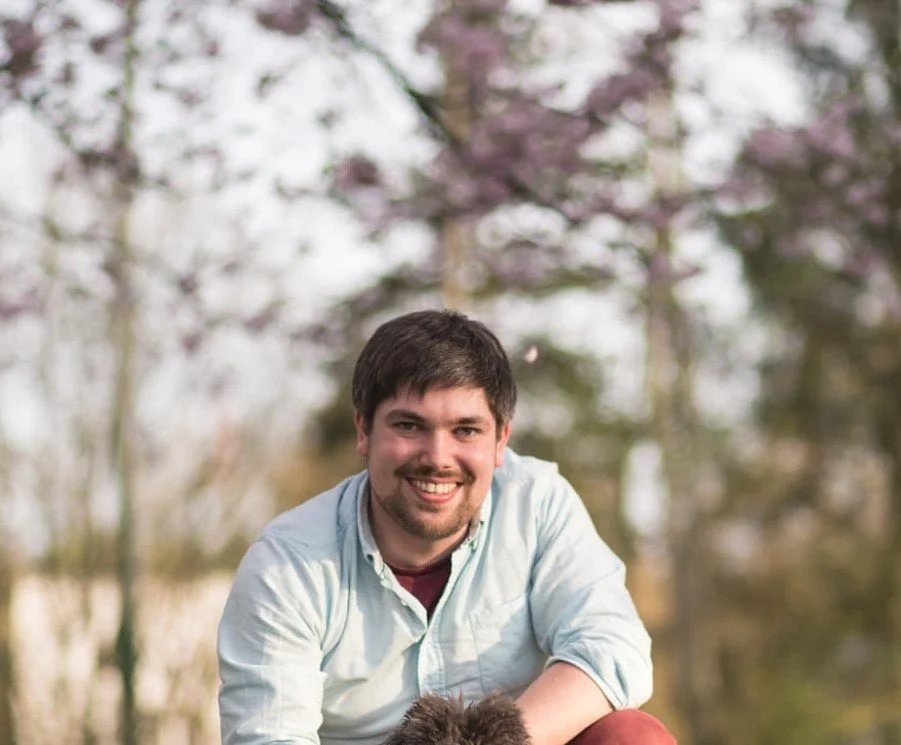A smiling man with dark hair and a beard outdoors, with blurred trees and pink blossoms in the background.