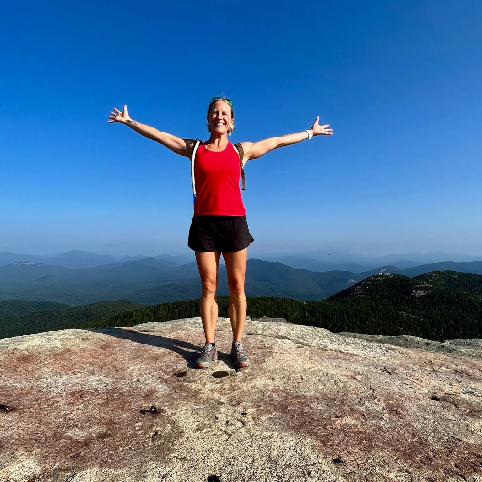 A woman standing on a rocky mountaintop with arms outstretched, smiling, wearing a red tank top, black shorts, hiking shoes, and carrying a backpack, with a scenic view of mountains and blue sky in the background.