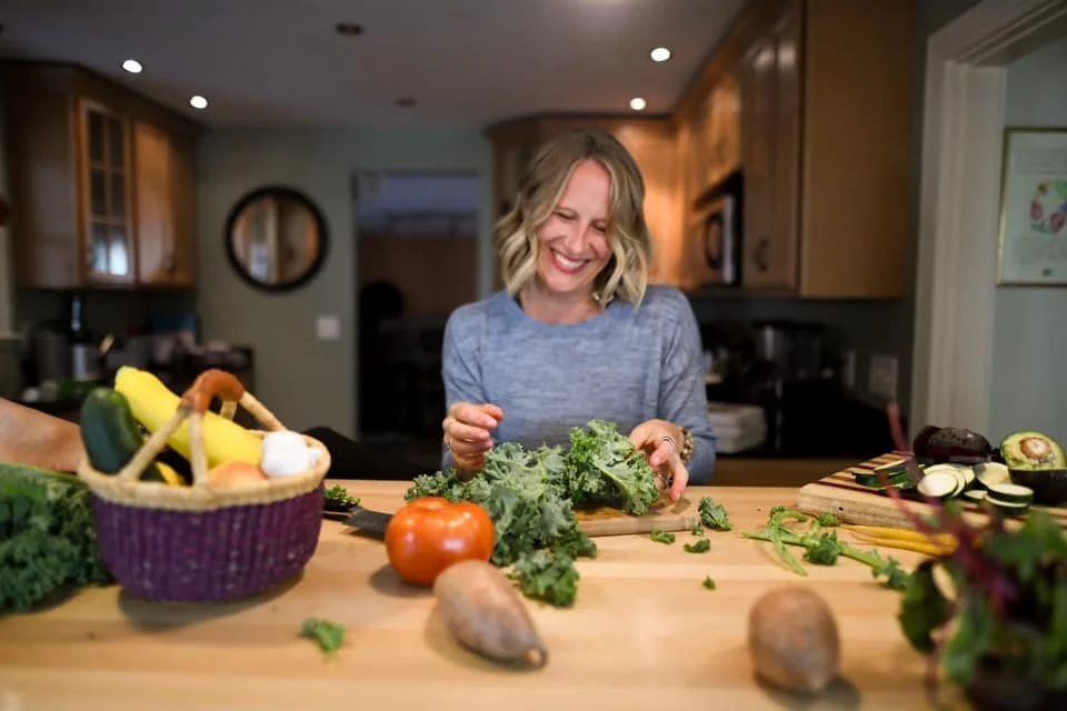 Woman chopping kale in a kitchen with various vegetables on the counter, including a tomato, potatoes, cucumbers, and a basket of bananas and other produce.
