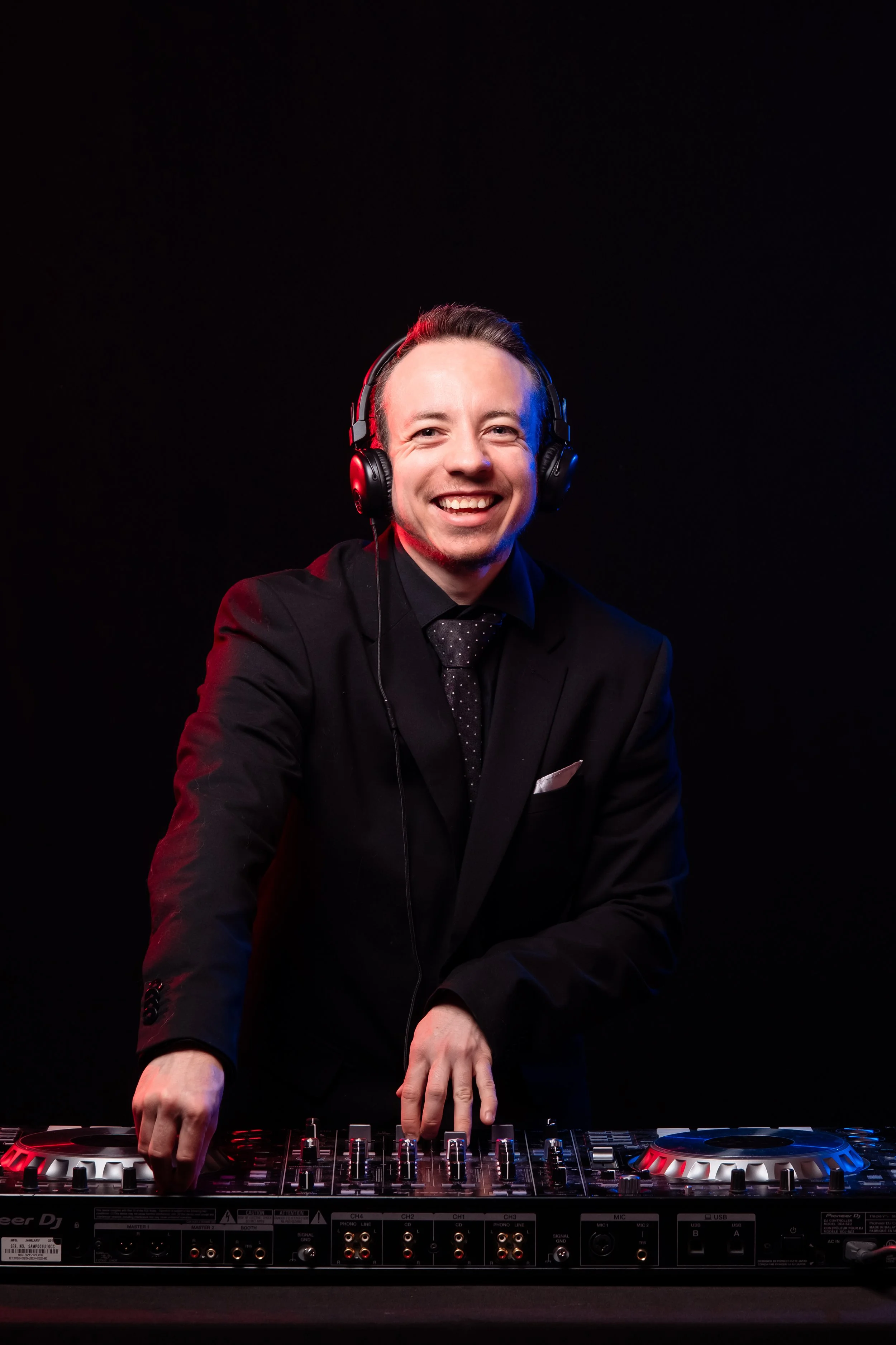A man in a black suit, black shirt, and dotted tie wearing headphones, smiling, standing behind DJ equipment in a dark studio with red and blue lighting.