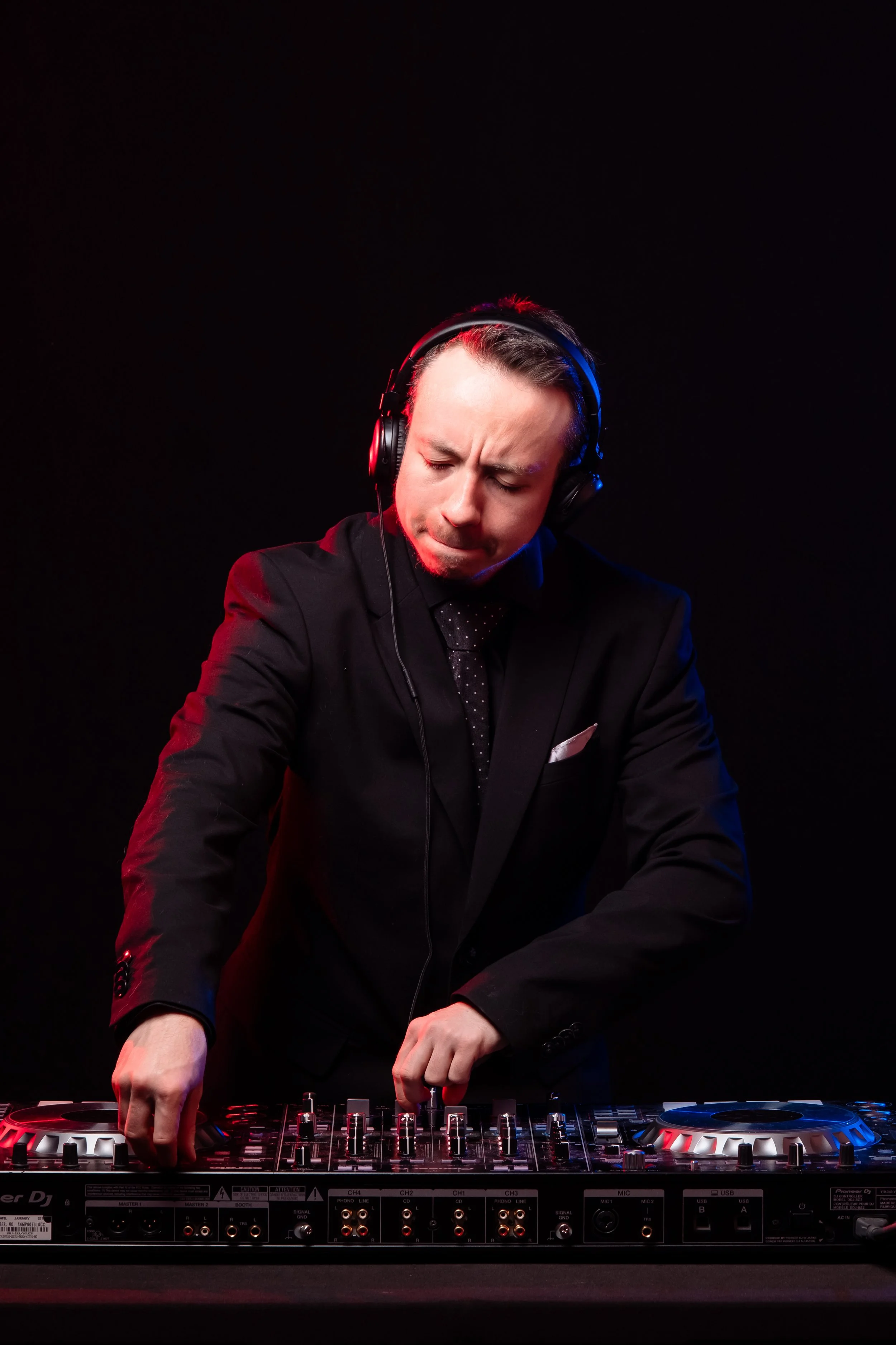 A man in a black suit with a polka dot tie and white pocket square DJing with headphones, focused on the mixing equipment against a black background.
