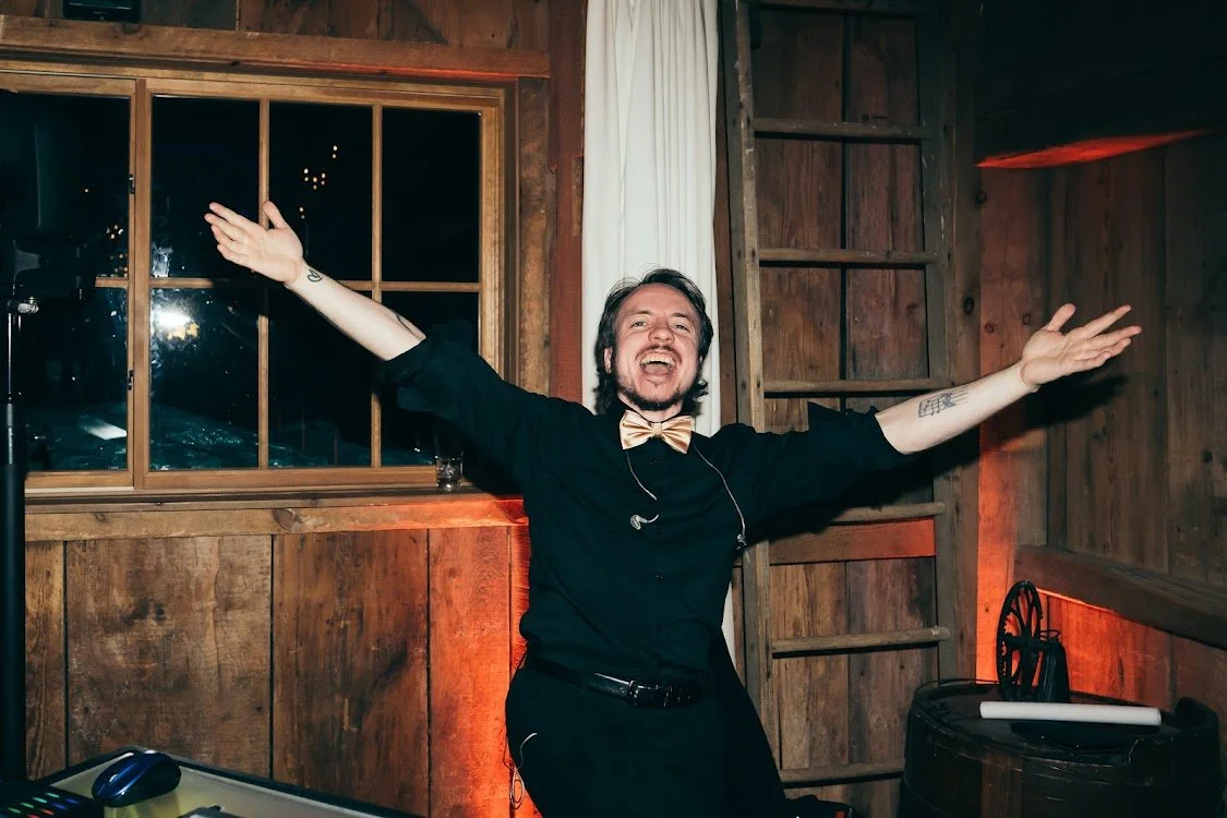 A man dressed in black with a gold bow tie, smiling with arms outstretched, standing in a rustic wooden room.