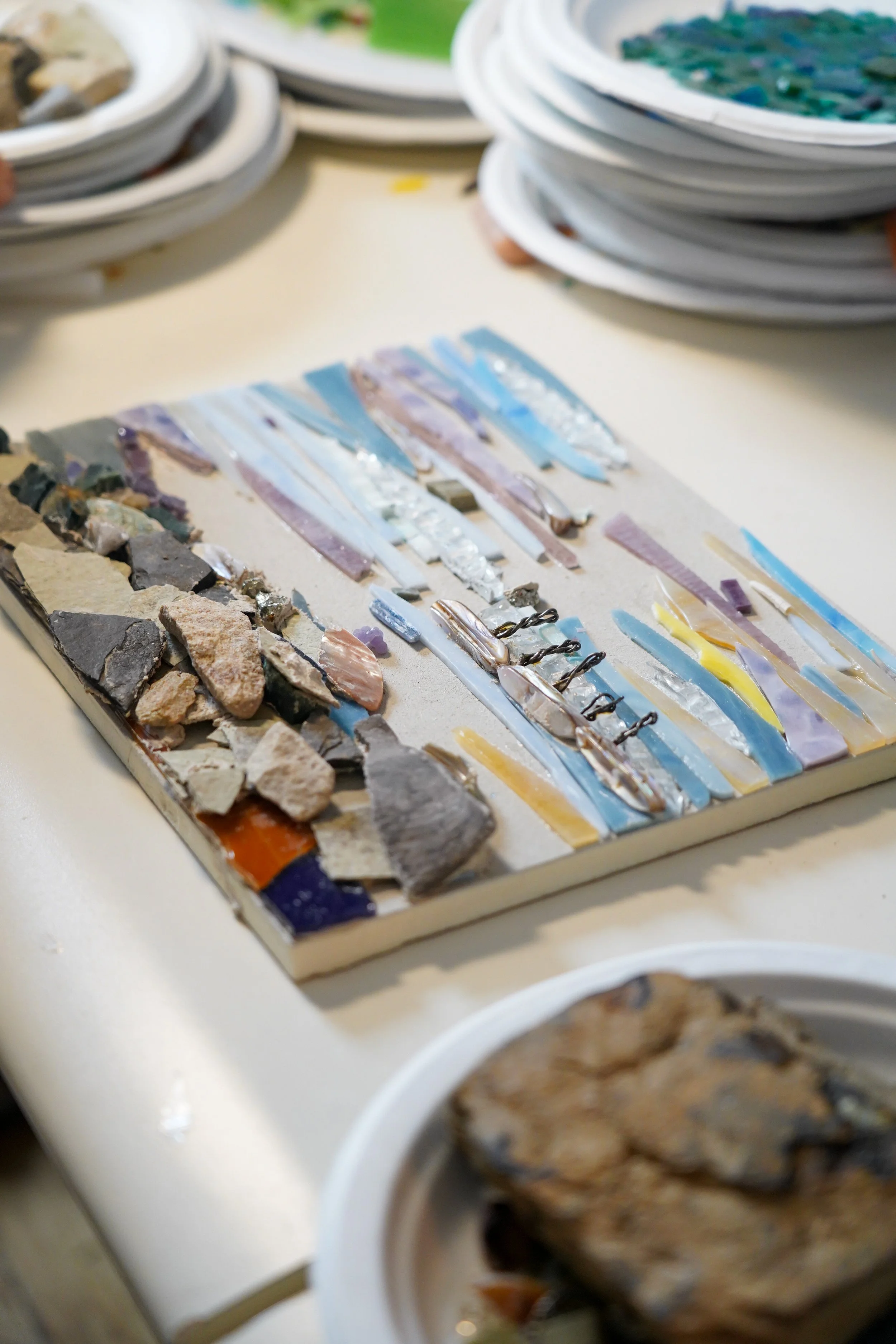 Mosaic art supplies with strips of colored glass and stones on a cardboard base on a table, with stacked plates in the background.