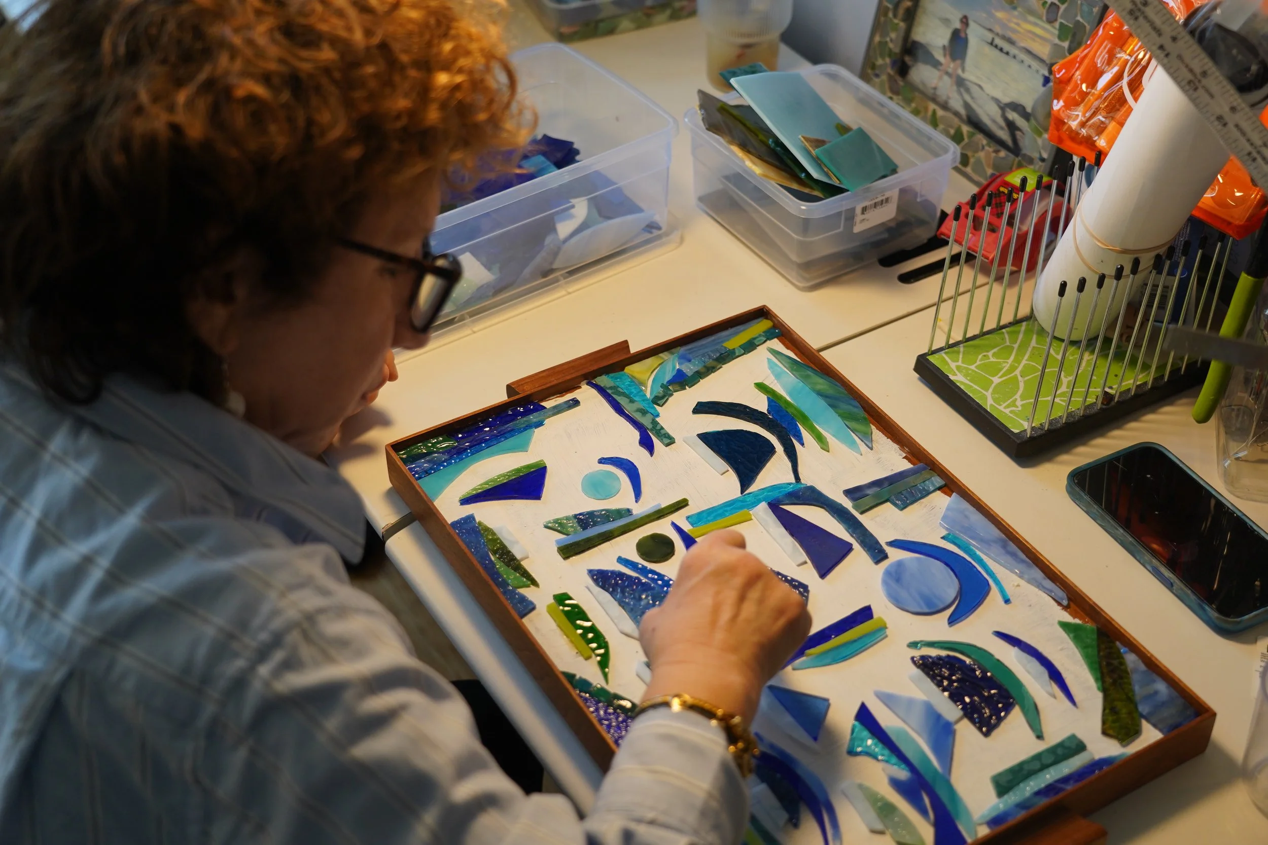 A woman with curly red hair and glasses working on a mosaic art piece at a cluttered workspace, surrounded by various tools and supplies.