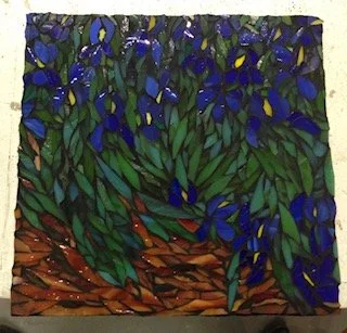 A stained glass artwork depicting irises, leafy green and blue foliage with a brown background.