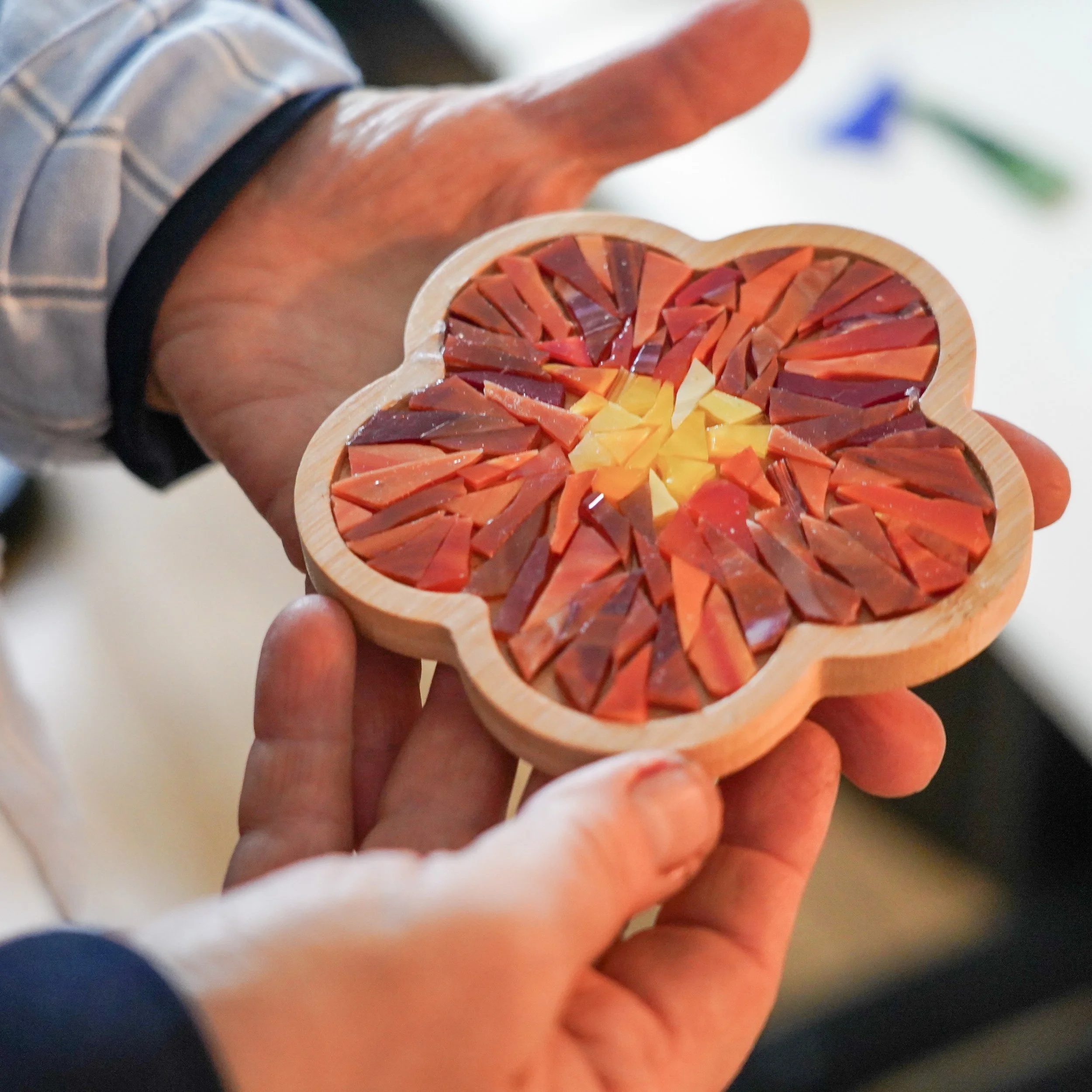 A person holding a wooden flower-shaped coaster filled with cut slices of red and yellow glass.