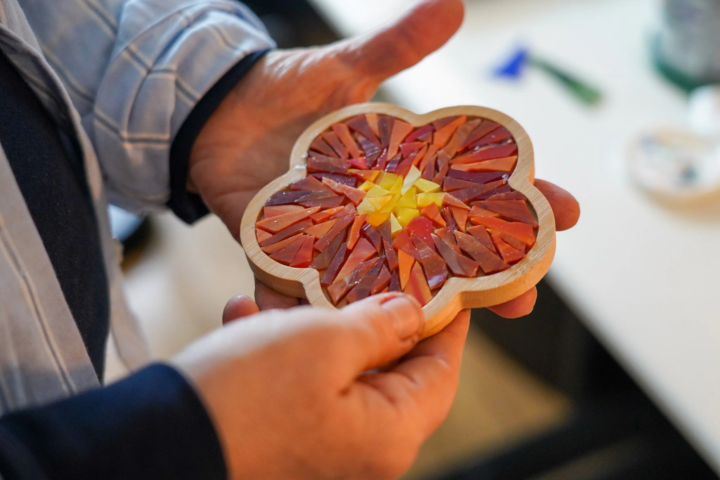 Hands holding a flower-shaped wooden coaster filled with small, colorful pieces of glass or stone arranged in a mosaic pattern.