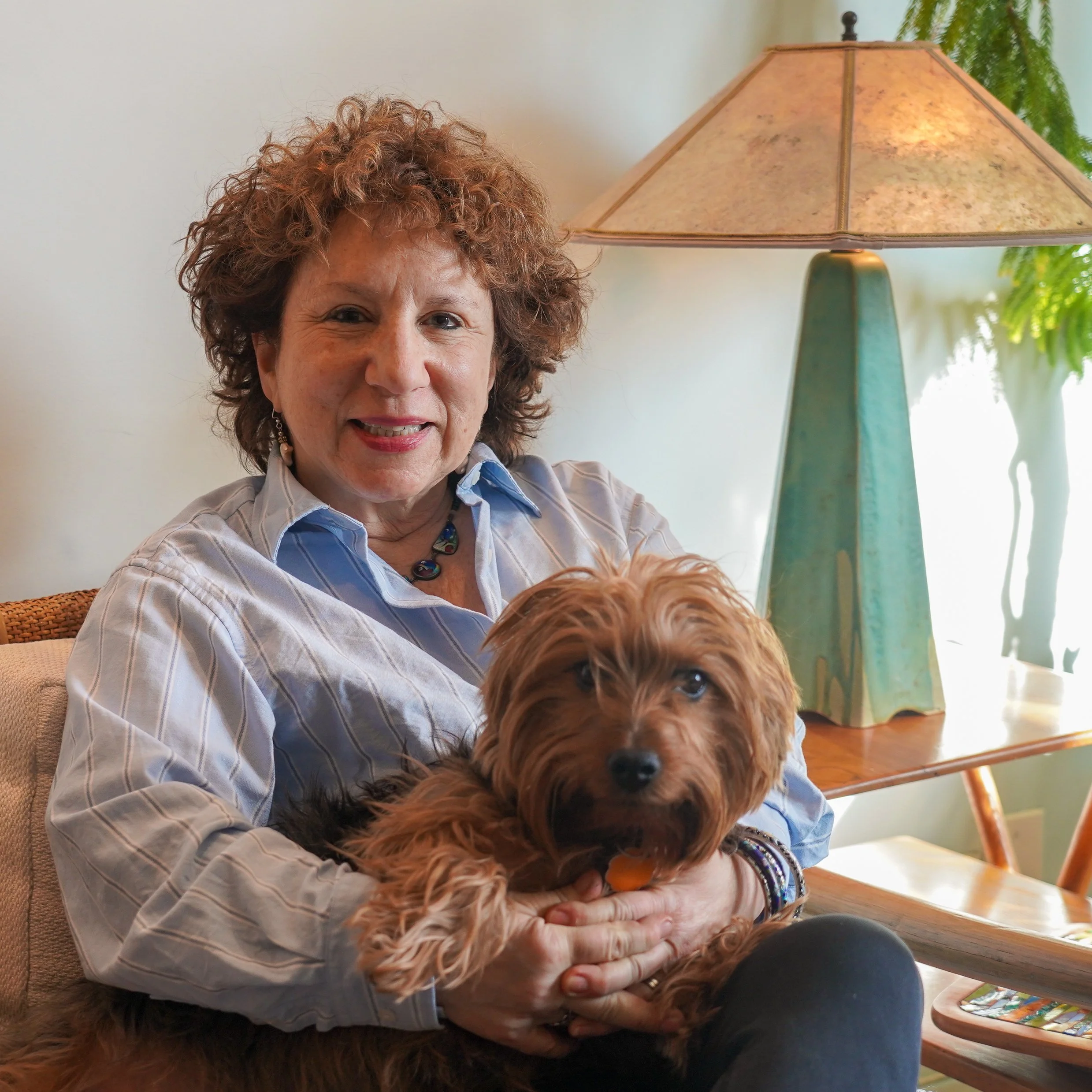 A woman with curly hair smiling, sitting on a beige sofa, holding a small brown dog with long ears. There is a green ceramic lamp with a beige shade on a wooden table behind her and a potted plant nearby.