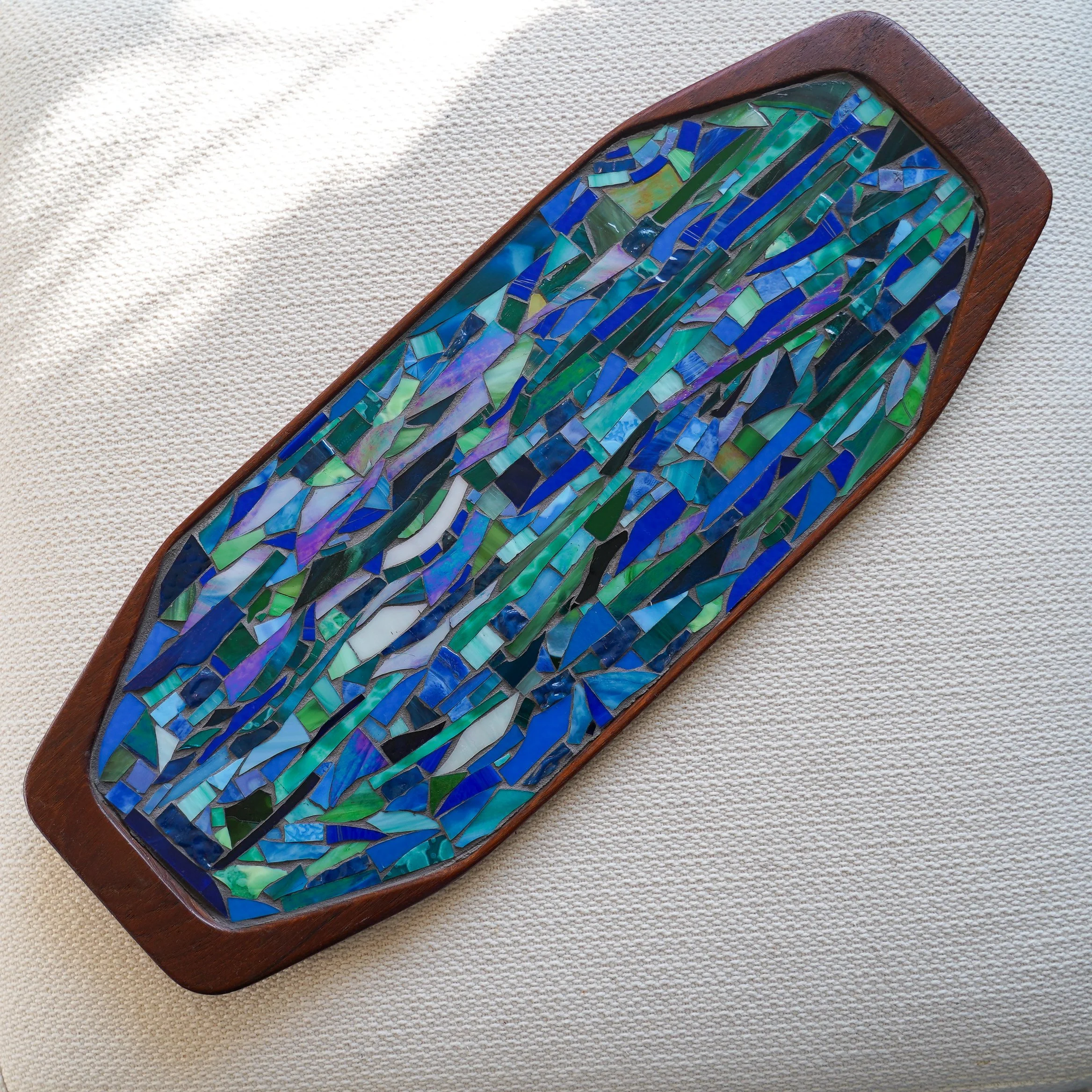 A rectangular mosaic with blue and green glass pieces in a wooden frame, placed on a textured beige fabric surface.