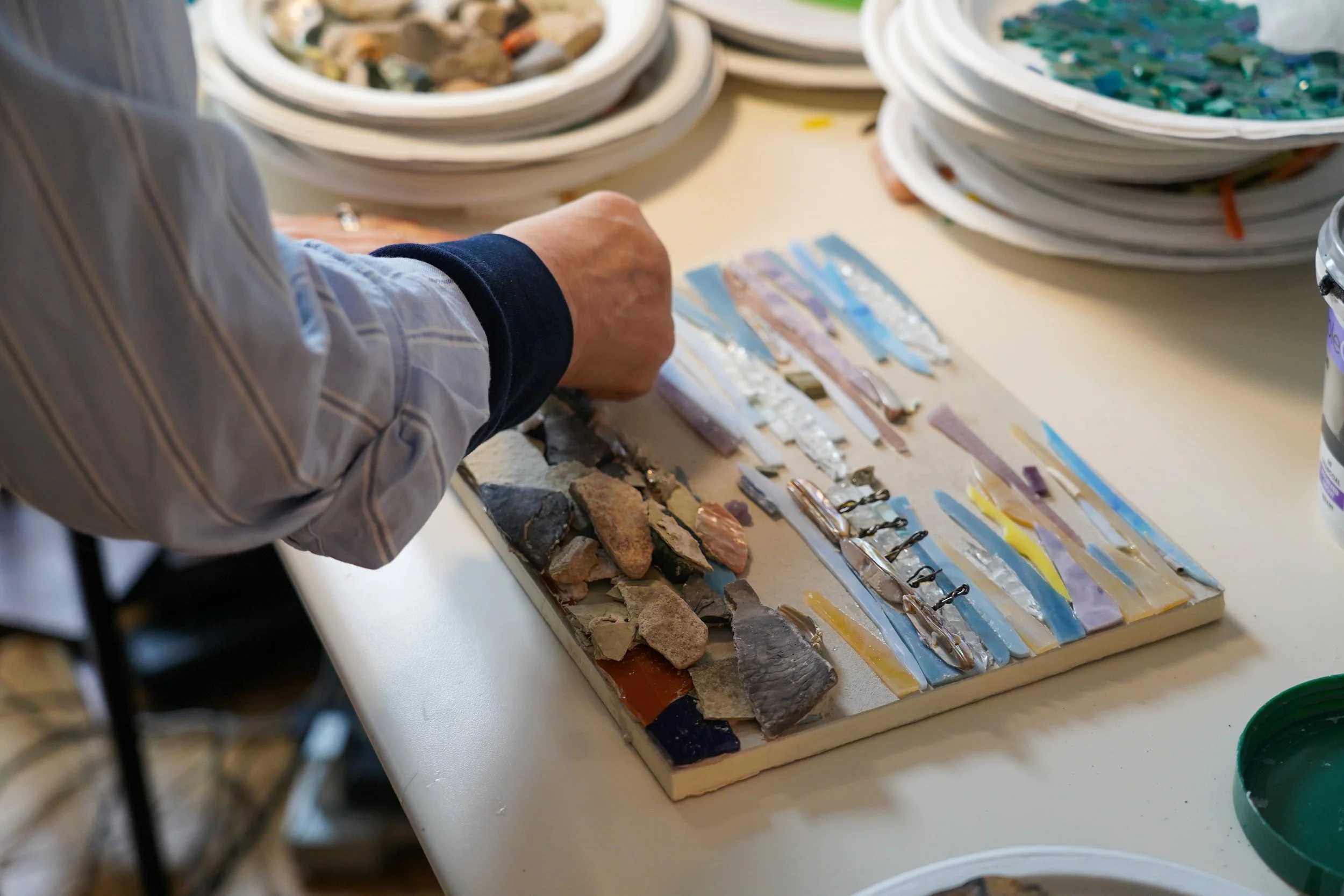 A person's hand arranging colorful broken glass pieces and rocks on a display board, with plates of mosaics and mosaic materials in the background.