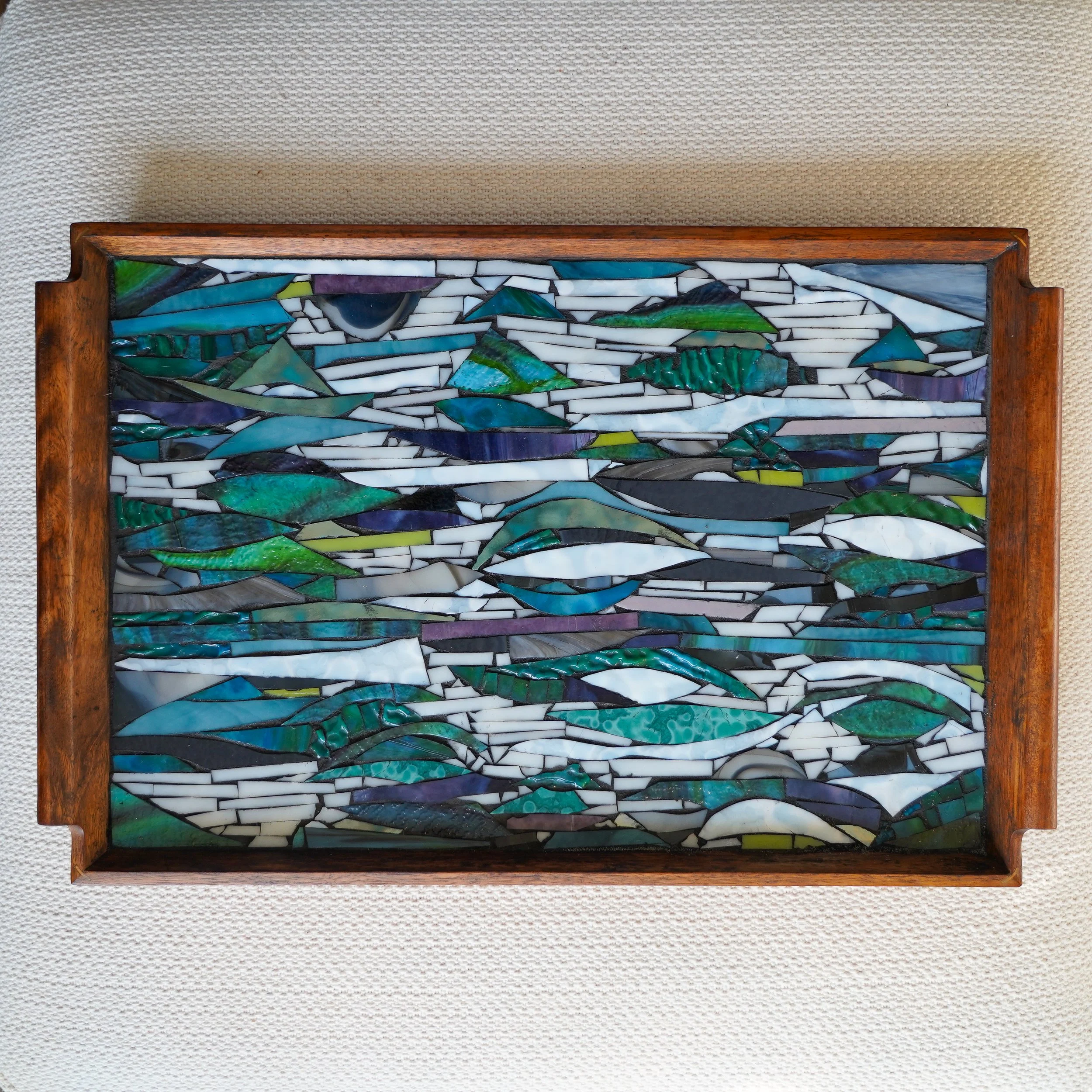 A mosaic art piece with various shades of green, blue, white, purple, and black glass shards arranged in an abstract pattern, framed in a wooden border.