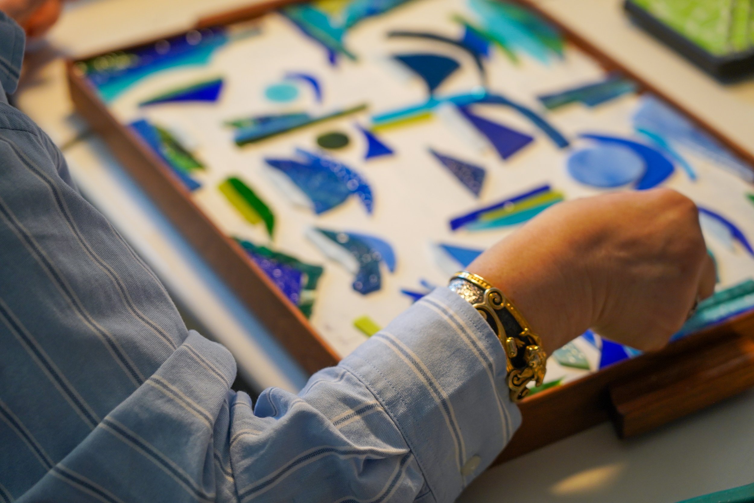 Person working on a mosaic artwork with blue and green glass pieces in a wooden tray, wearing a striped shirt and decorative bracelets.