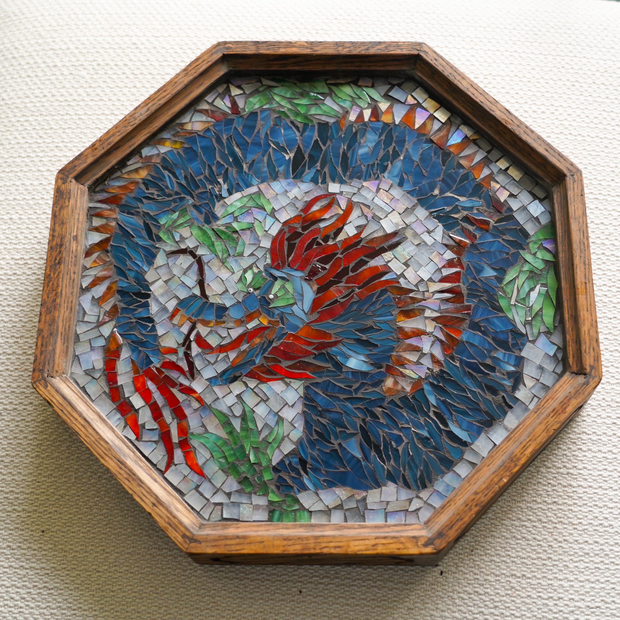 An octagonal wooden frame containing a colorful mosaic of a dragon, with deep blue, red, green, and white tiles arranged to depict the bird's feathers and body, on a light fabric background.