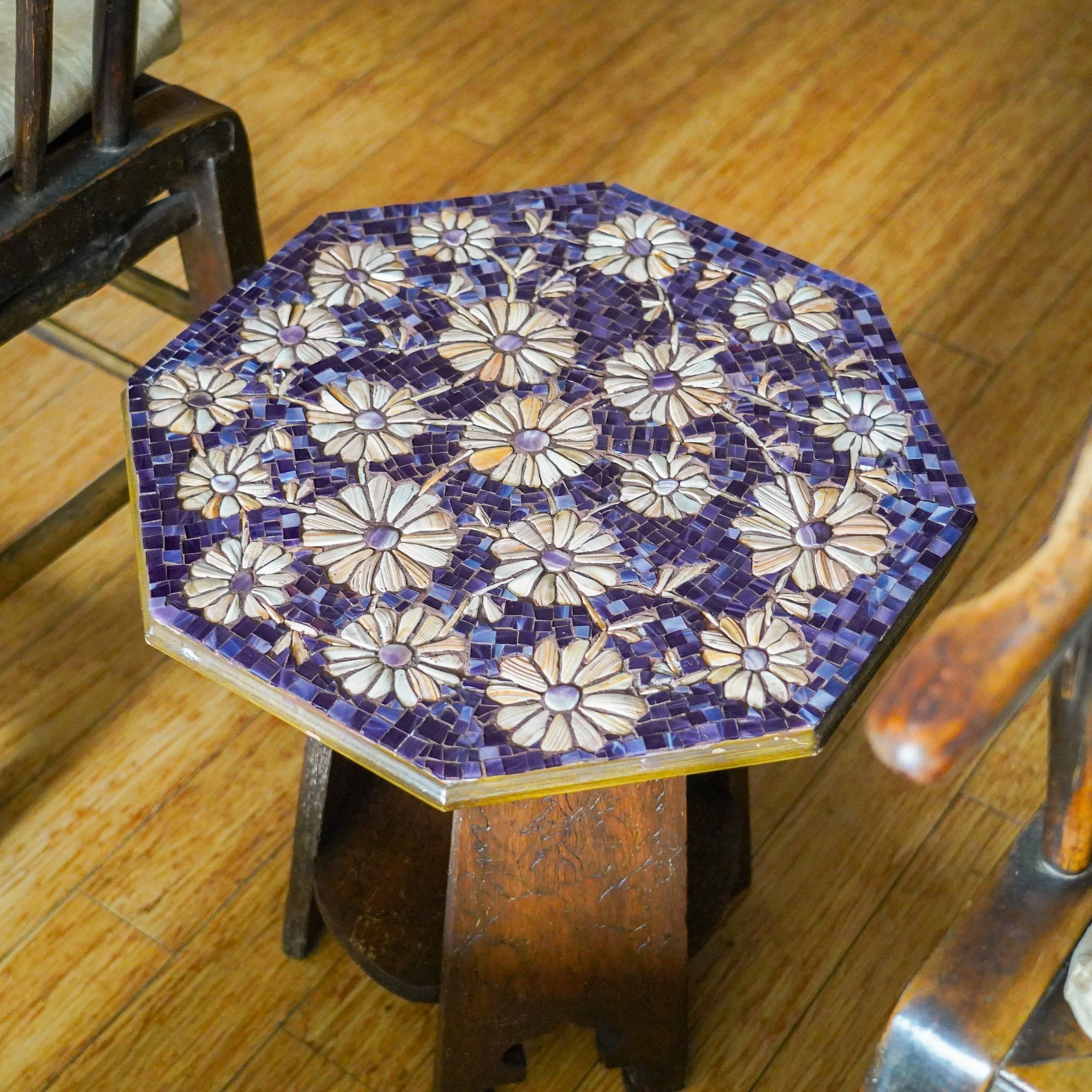 Hexagonal table with a mosaic top featuring white and purple daisy flower patterns, surrounded by wooden chairs with a wooden floor.