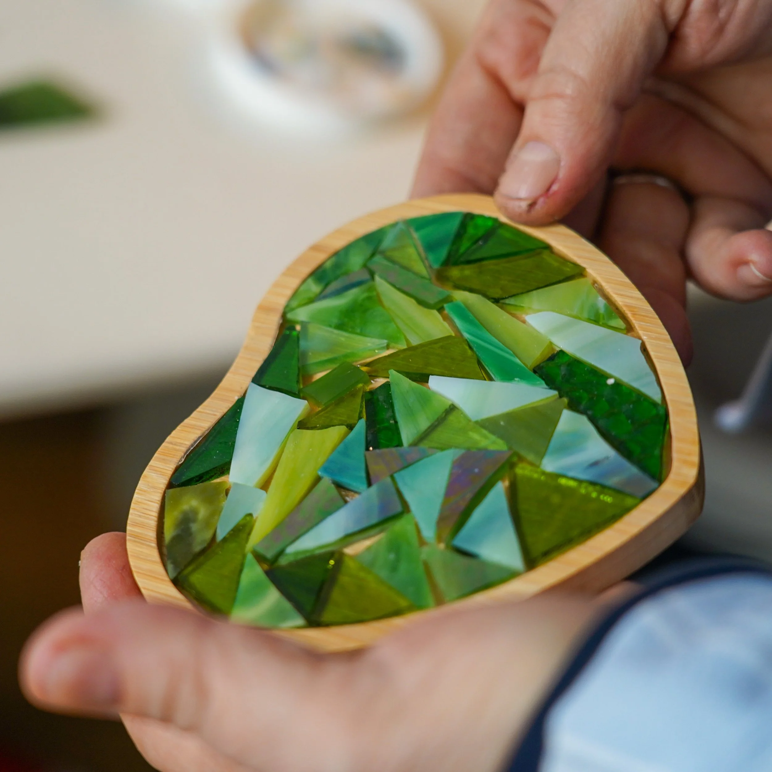 Person holding a wooden coaster with an assortment of small, irregularly shaped green and blue glass fragments.