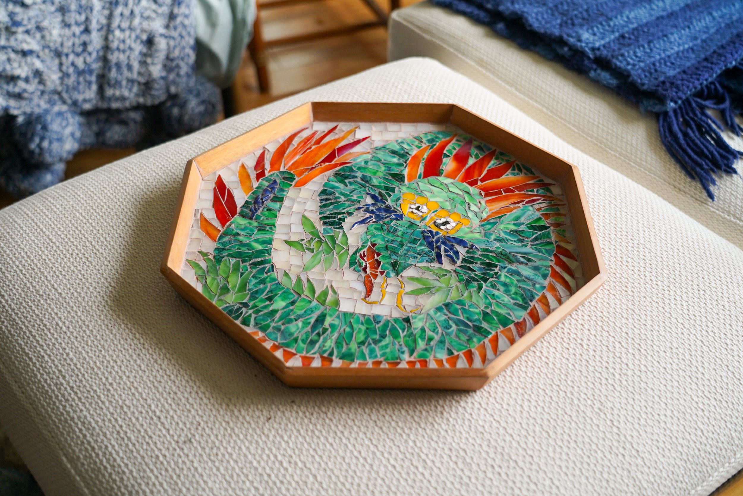 A mosaic artwork of a colorful dragon inside a wooden frame, resting on a beige fabric surface.