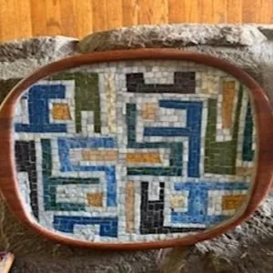 Close-up of an oval tray with a brown frame, reflecting a mosaic wall with blue, white, black, yellow, and green tiles in a geometric pattern.