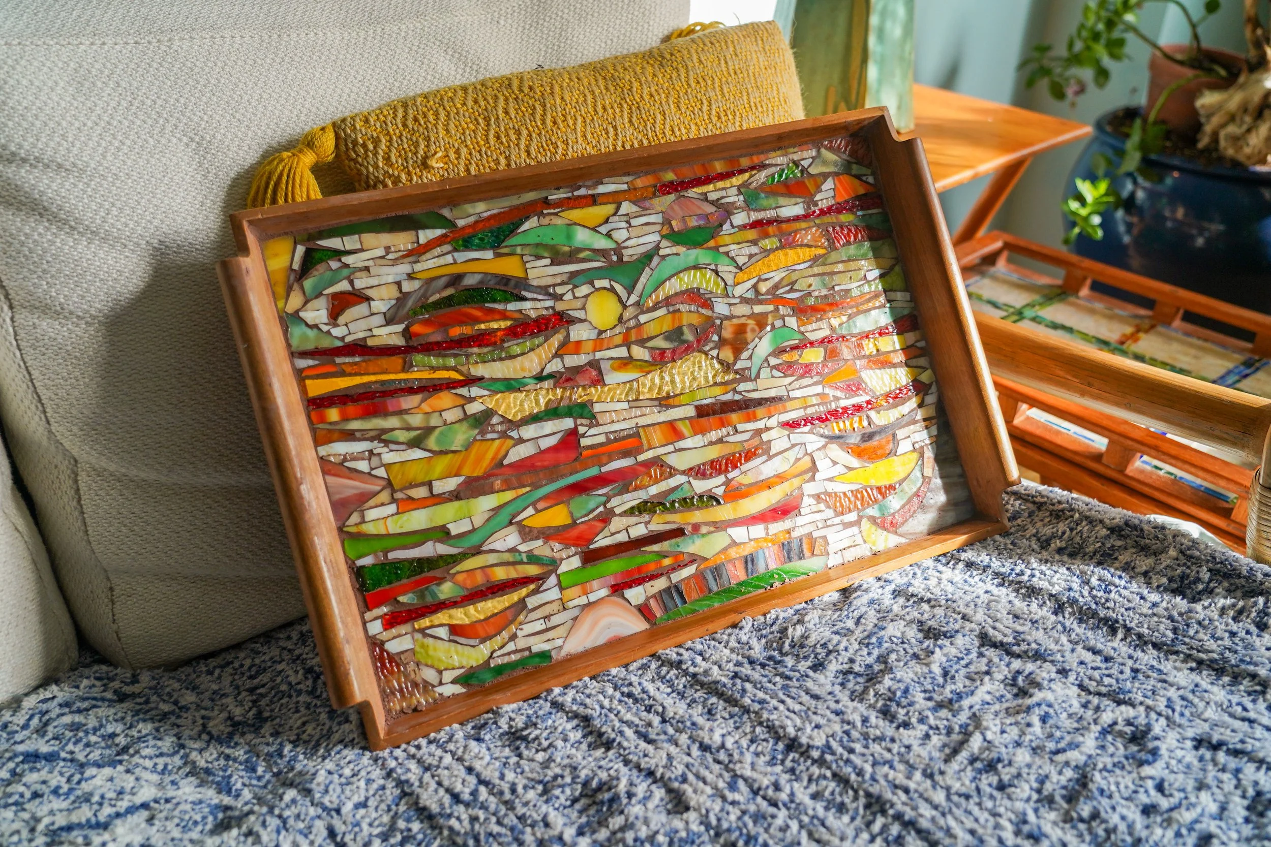 A mosaic art piece with colorful glass pieces in a wooden frame, resting against a beige cushion on a sofa.