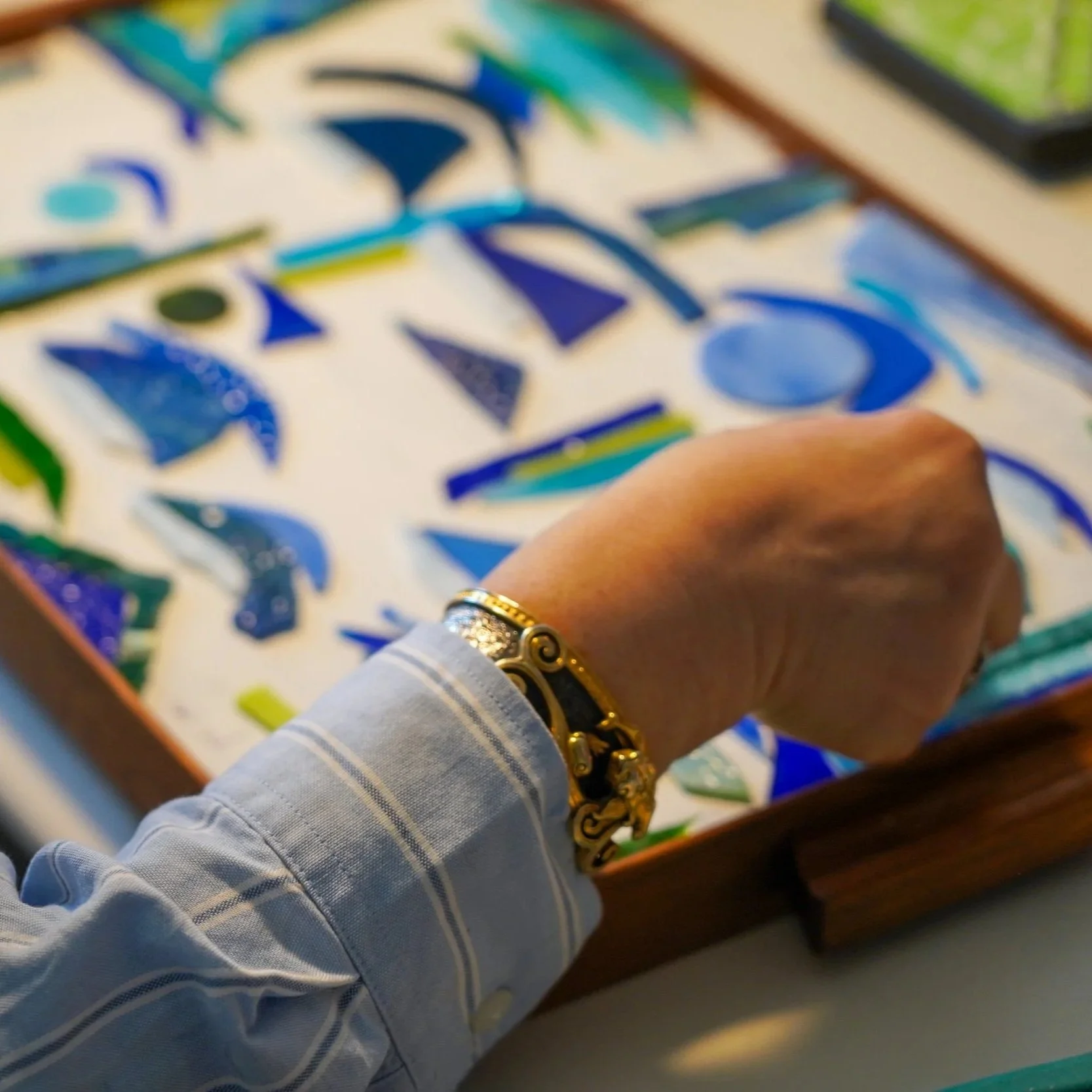 A person's hand and wrist adorned with gold and silver jewelry are positioned over a tray of various colorful, abstract jewelry pieces.
