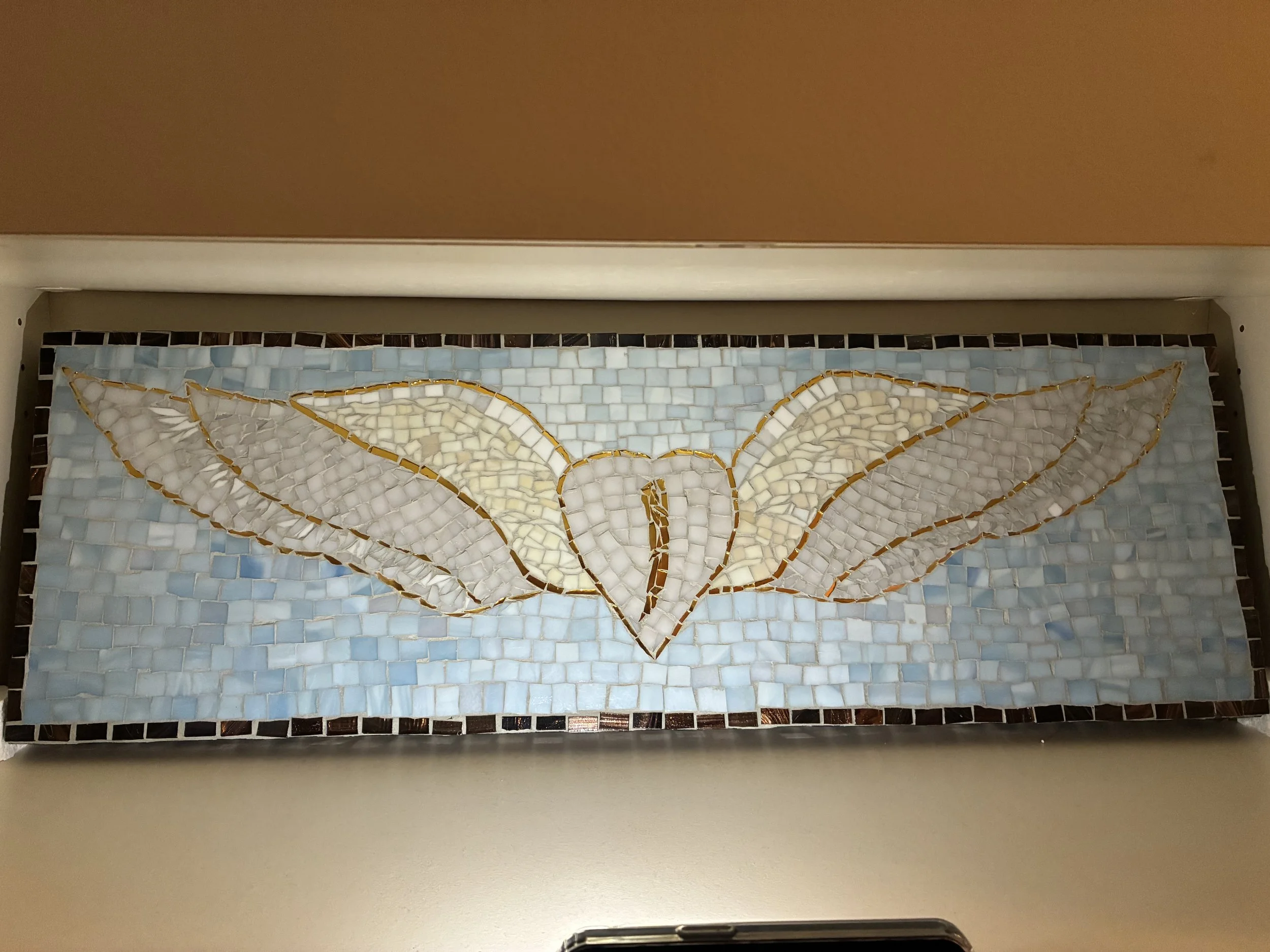 A mosaic artwork featuring a white heart with white wings outlined in gold, set against a light blue background.