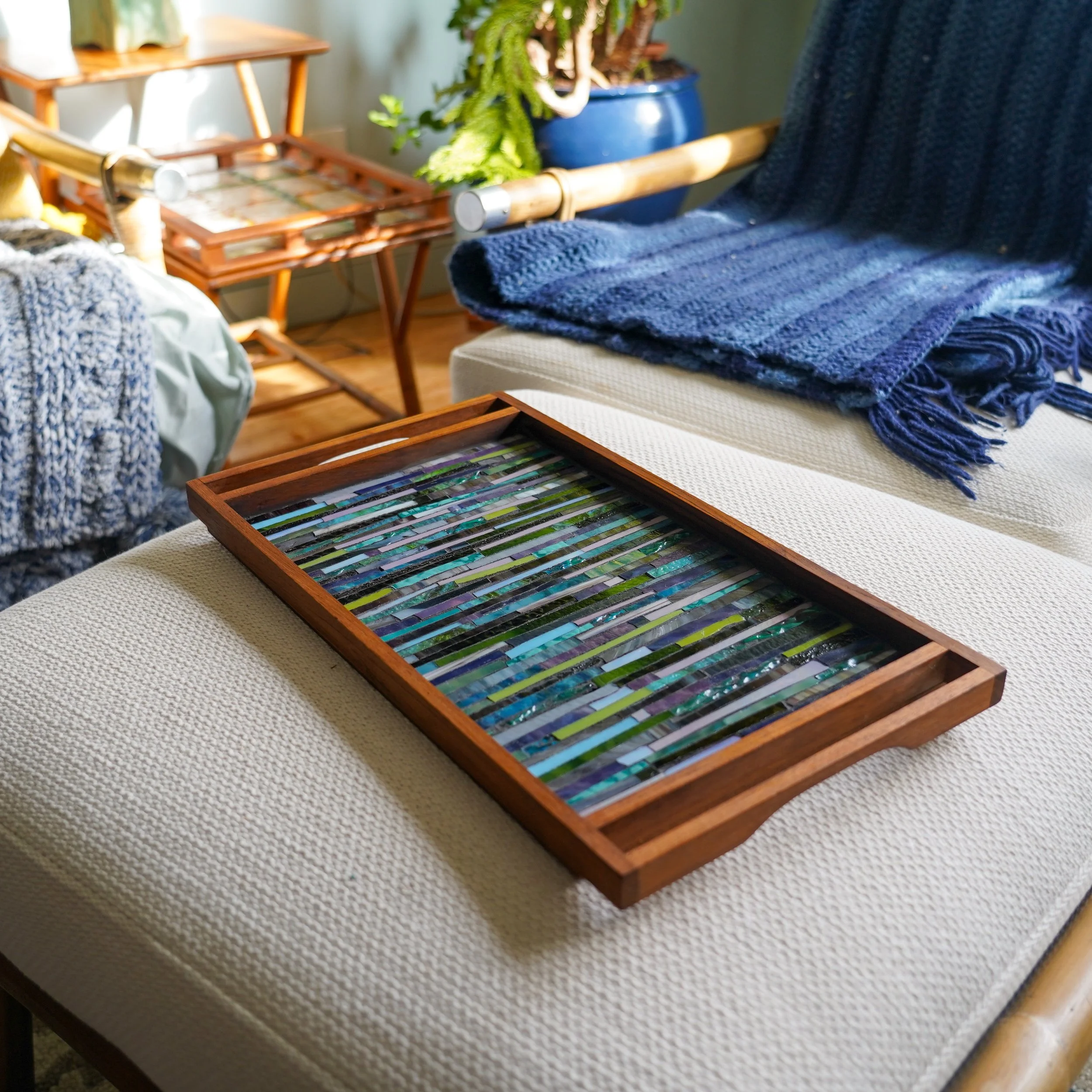 A wooden tray with colorful glass mosaic strips rests on a beige textured cushion, with a dark blue knitted blanket draped over a chair nearby. In the background, there are side tables, a potted fern plant, and various decorative items.