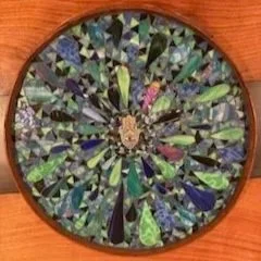 A circular mosaic with various green, blue, and pink glass pieces arranged in a radial pattern.