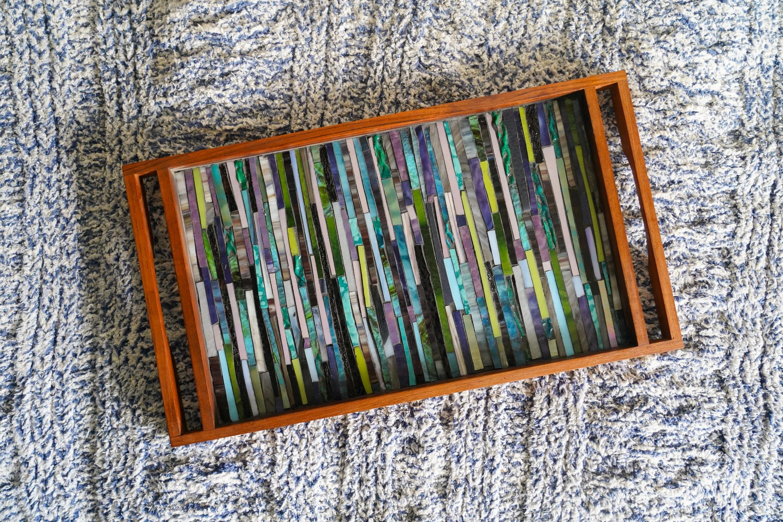 Wooden tray filled with multicolored glass mosaic tiles, placed on a plush gray and white textured blanket.