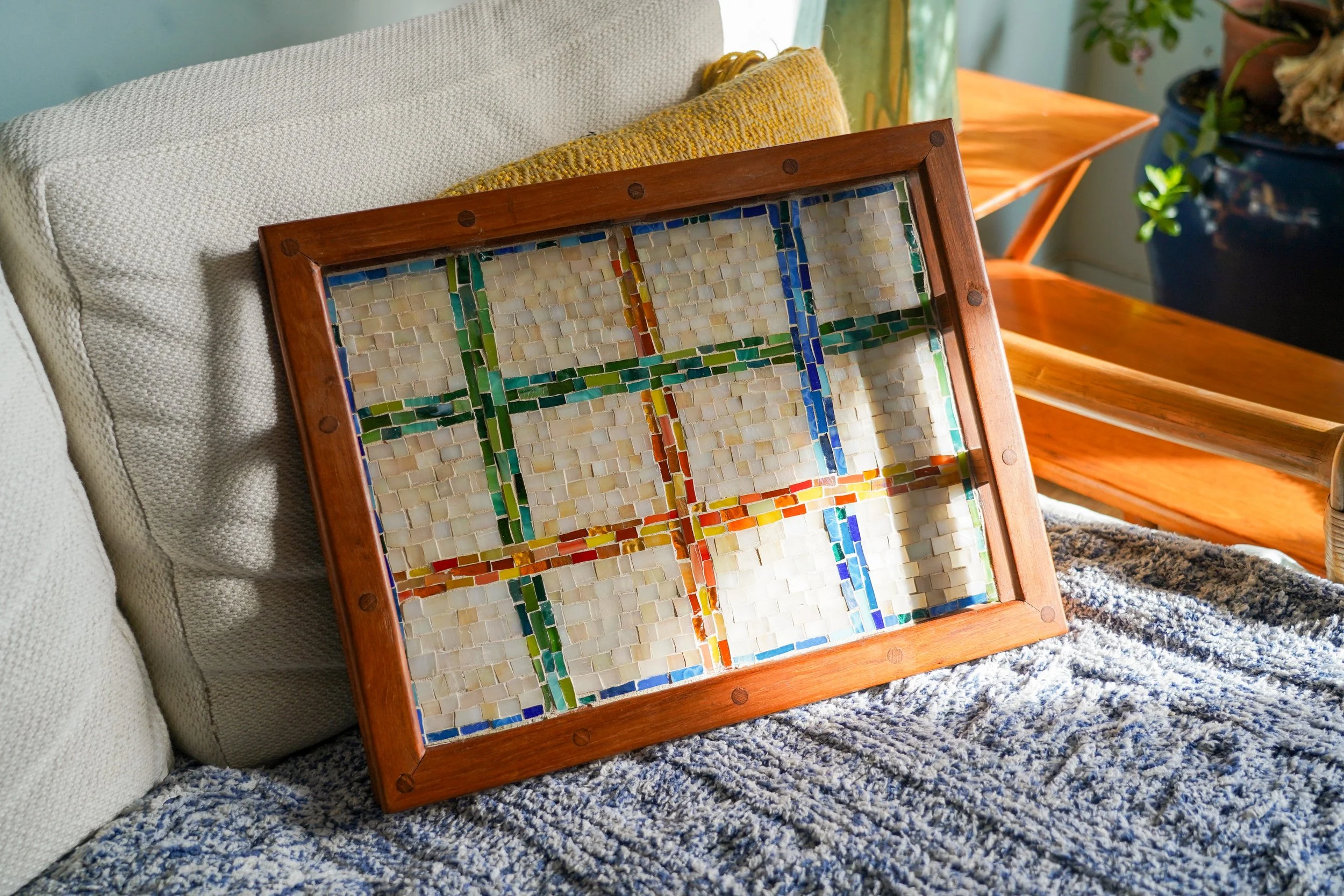 Colorful mosaic glass tray with a wooden frame resting on a beige couch next to yellow pillow and blue textured blanket.