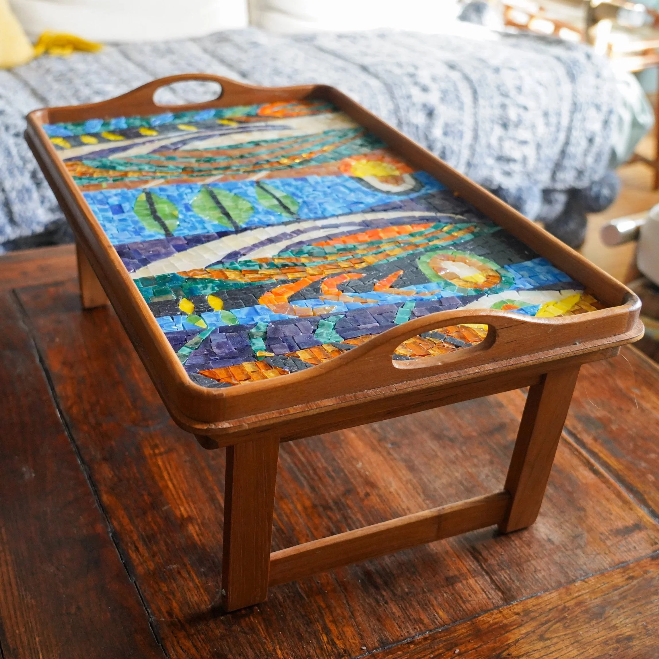 A wooden tray with a colorful mosaic design depicting a sunset over a lake with fish and lily pads, placed on a wooden table.