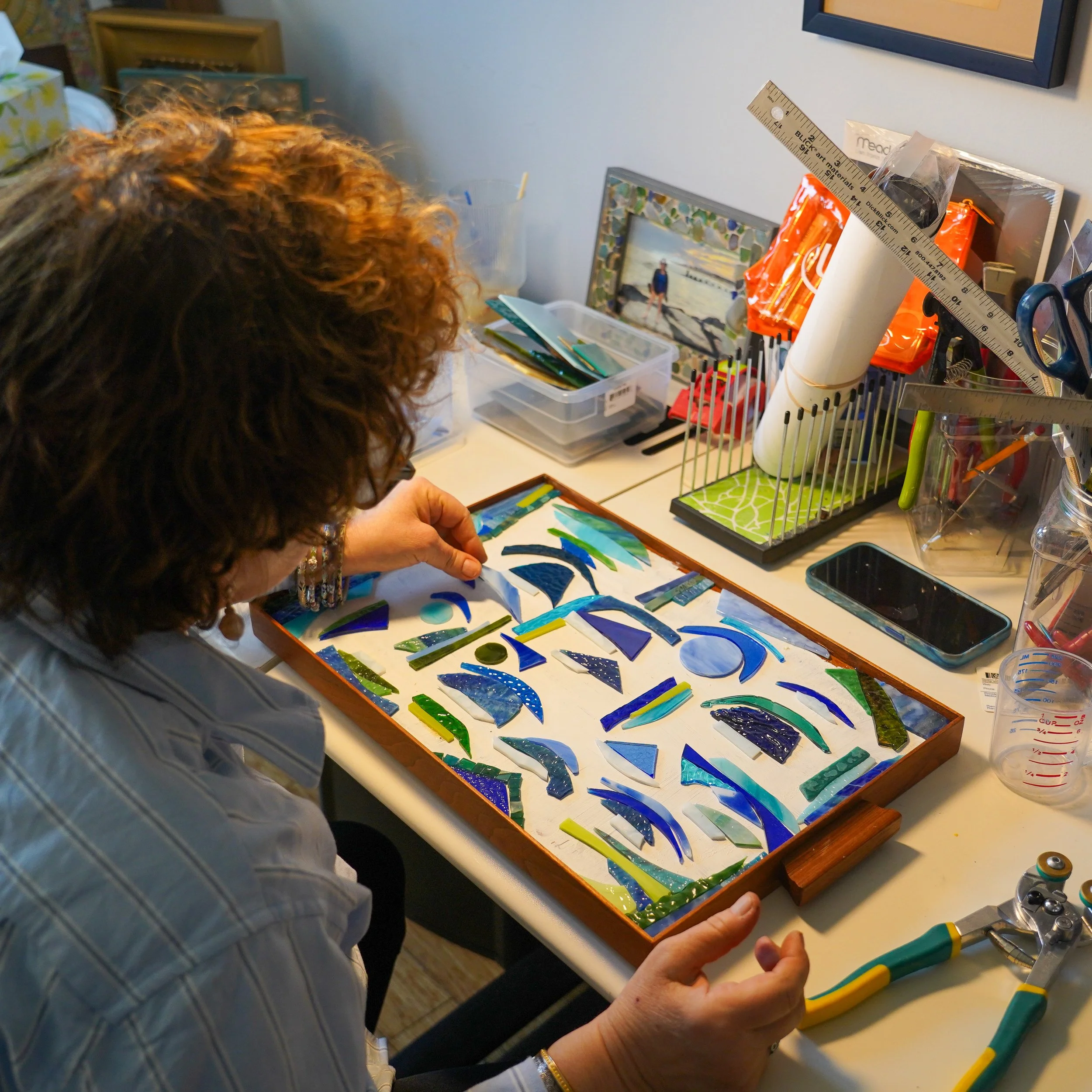 A person working on a mosaic art piece with colorful glass shards on a table in a craft room.