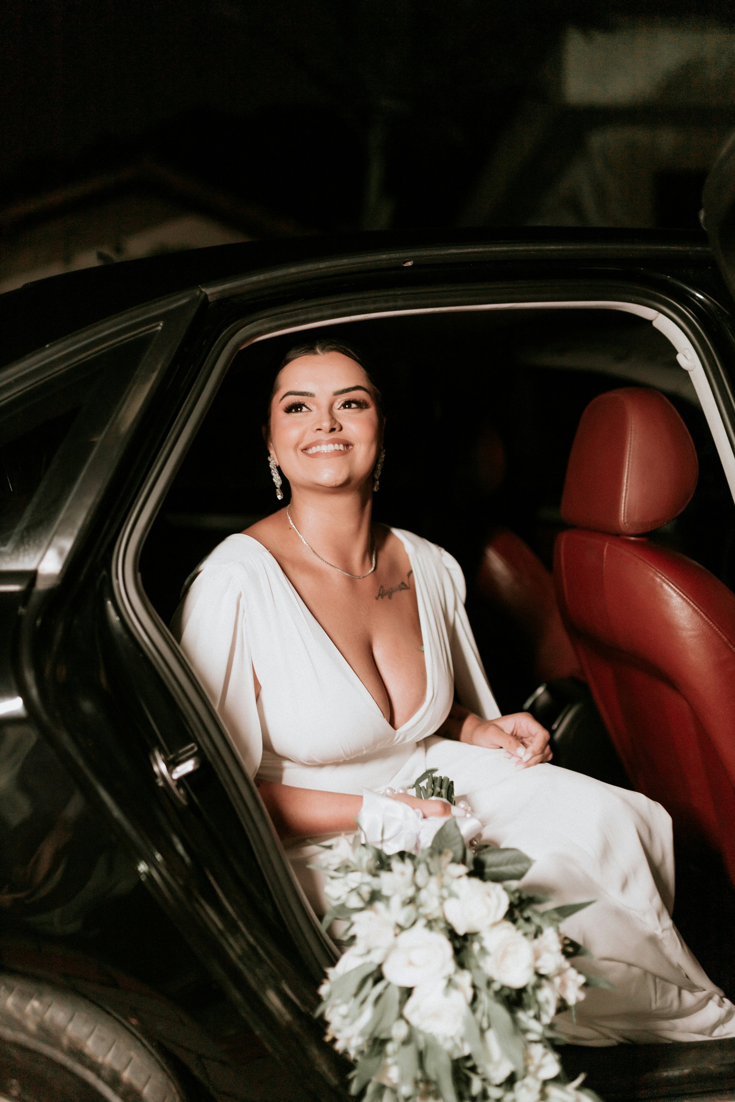 A smiling woman in a white dress sitting in a car, holding a bouquet of white flowers.